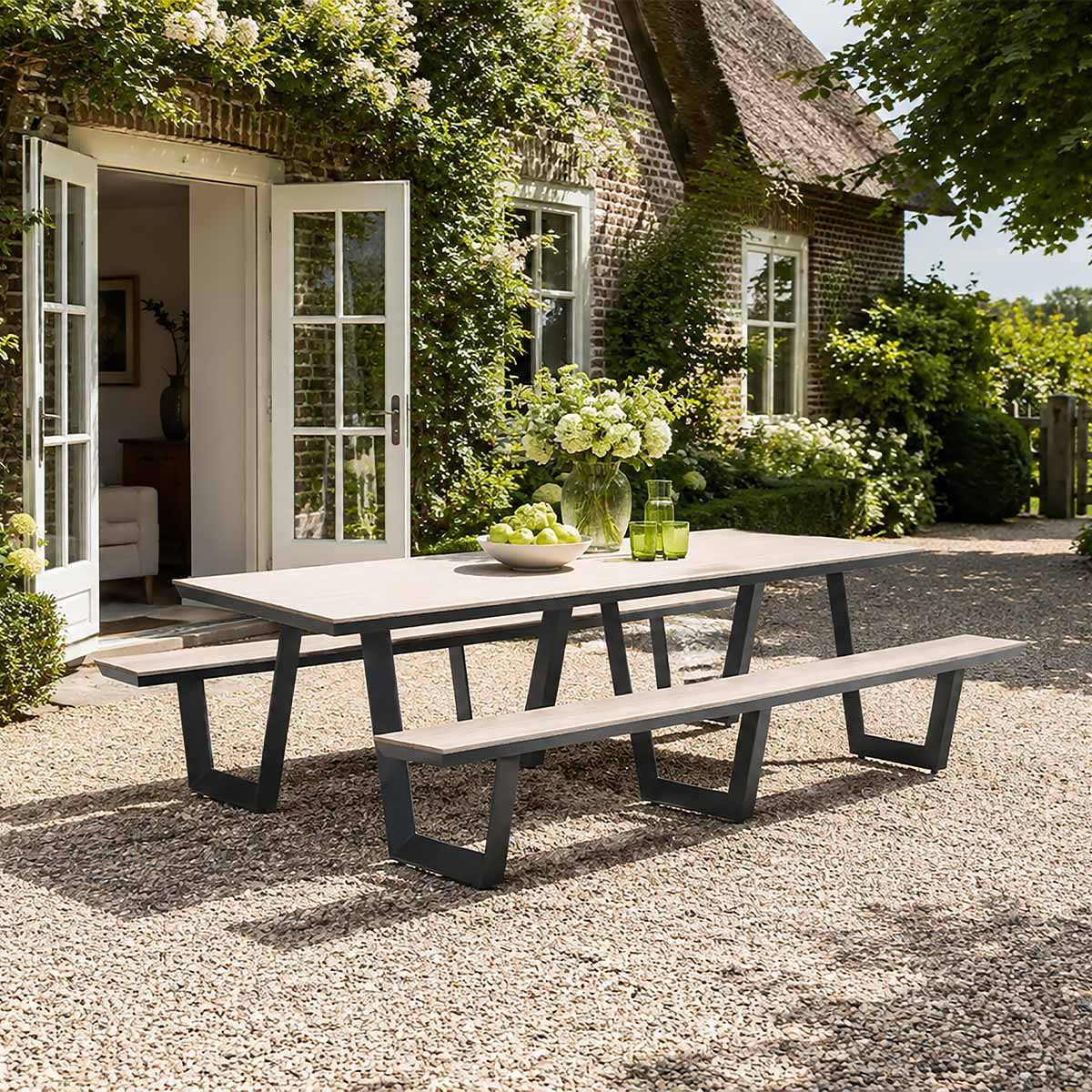 A rustic patio features a large wooden dining table with benches on gravel, set against a brick cottage with open doors.