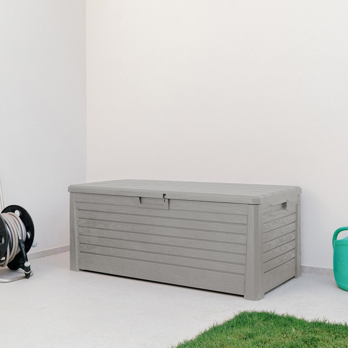 A grey storage box sits against a white wall outdoors, flanked by a hose reel and a green watering can.