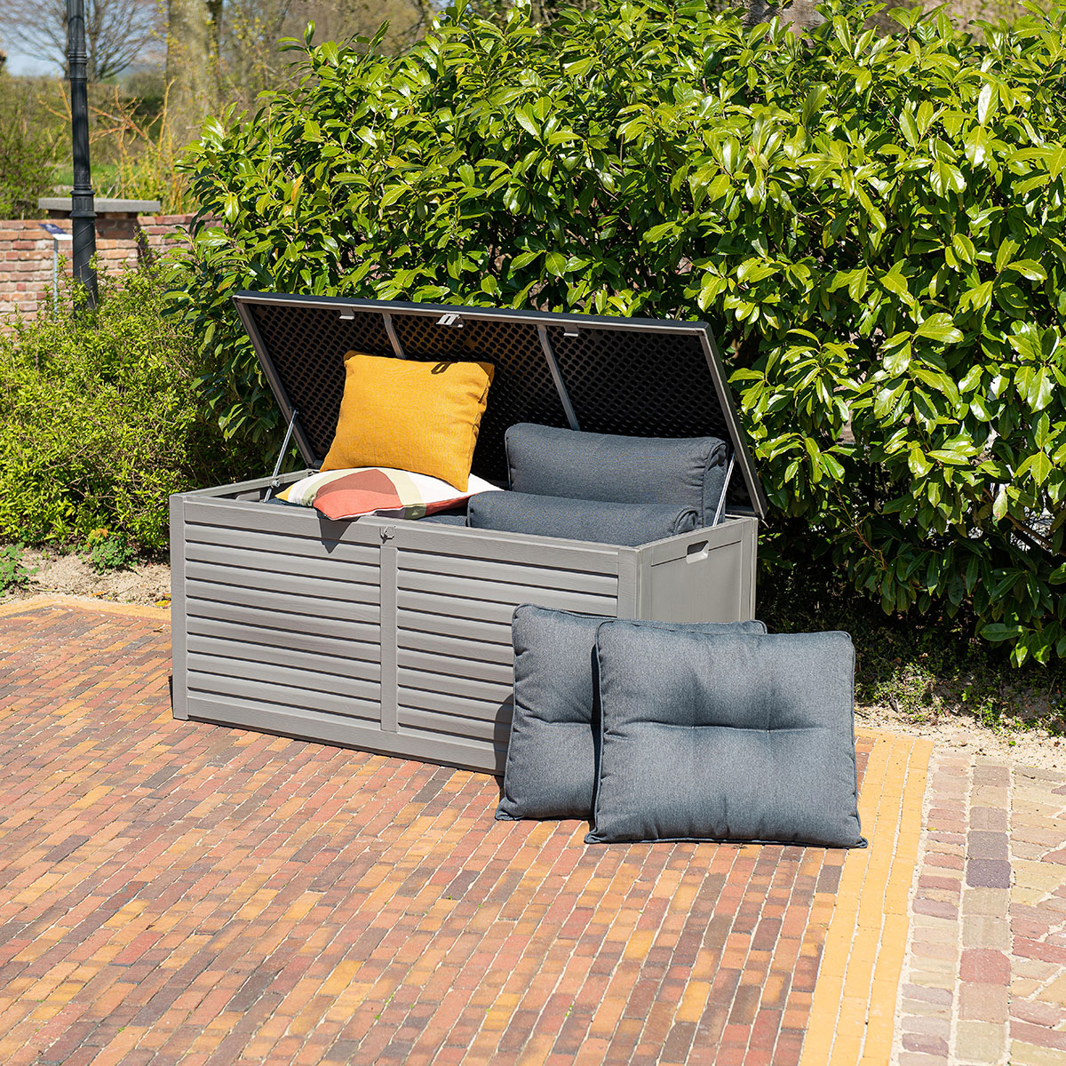 Grey garden storage box with open lid filled with cushions on red brick paving.