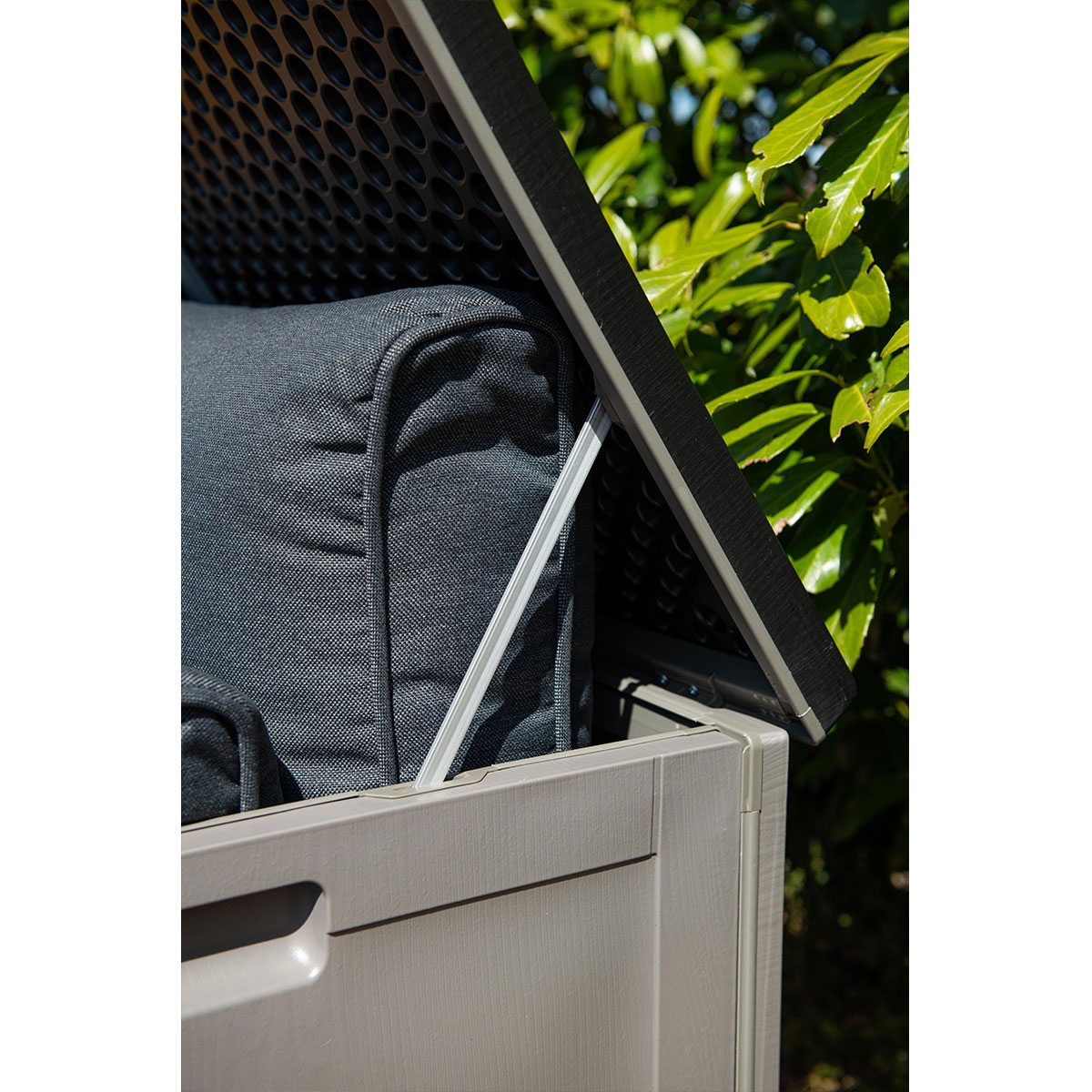 An open plastic storage box with a dark grey cushion inside, featuring a prop rod and lush green leaves in the background.