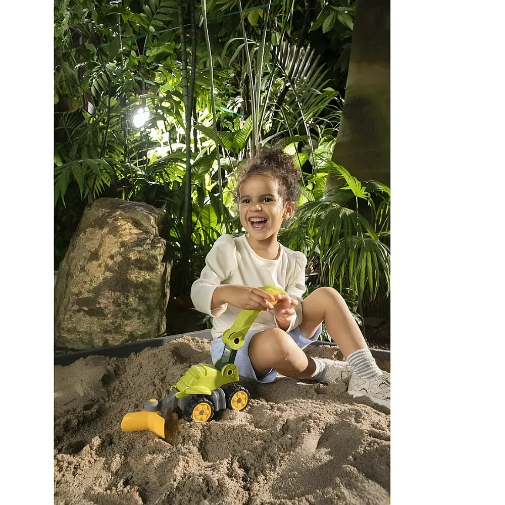 Ein glückliches Kind spielt im Sand mit einem grünen Bagger vor einer grünen Dschungel-Kulisse.