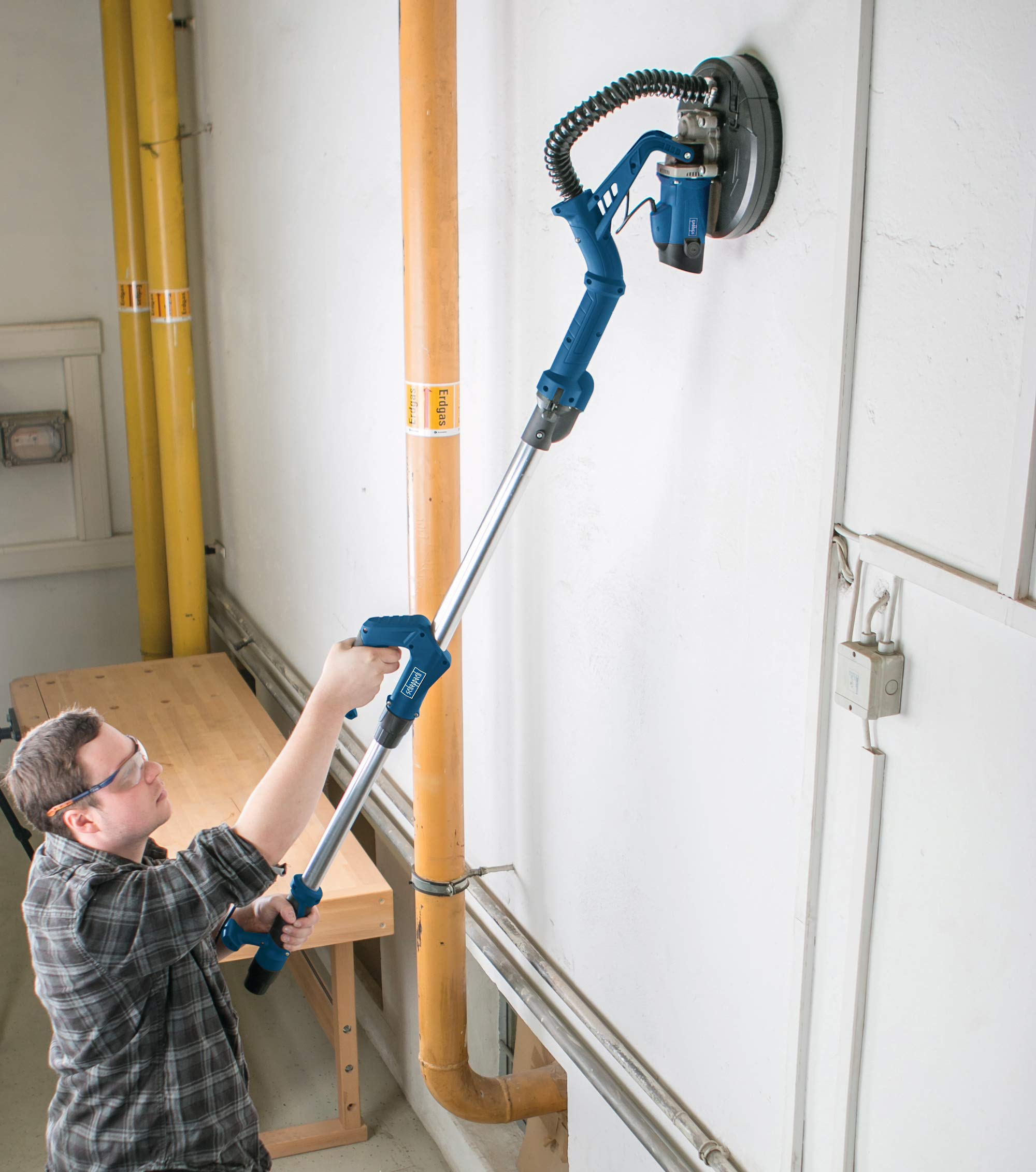Ein Arbeiter in Schutzbrille verwendet eine lange Schleifmaschine, um eine weiße Wand zu glätten.