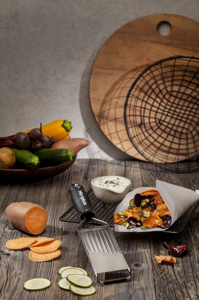 Eine Reibe liegt auf einem Holztisch neben Gemüseschips, frischem Gemüse und einer Schüssel mit Dip.