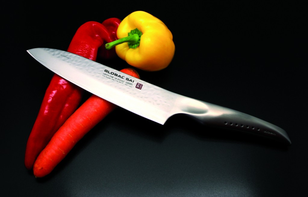 Ein Global Kochmesser liegt diagonal über roter Paprika und Karotte, mit gelber Paprika im Hintergrund.