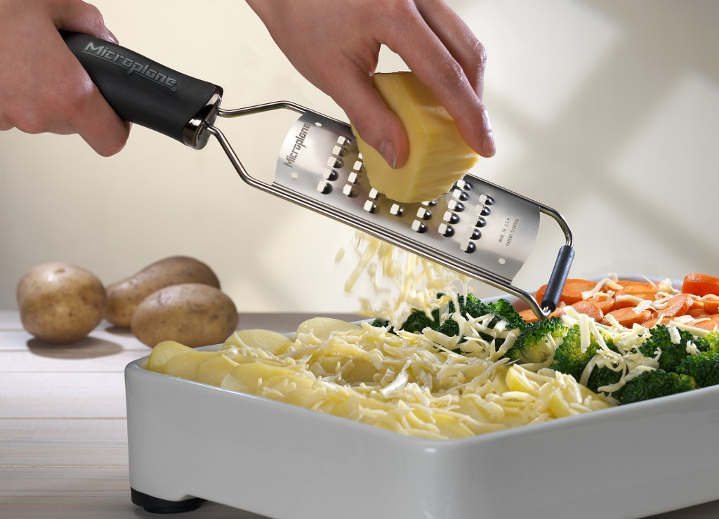 Eine Hand reibt Käse über einen Auflauf mit Kartoffeln, Brokkoli und Karotten.