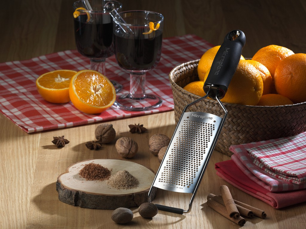 Eine Reibe, frische Orangen, Nüsse, Zimt und Glühwein auf einem Holztisch.