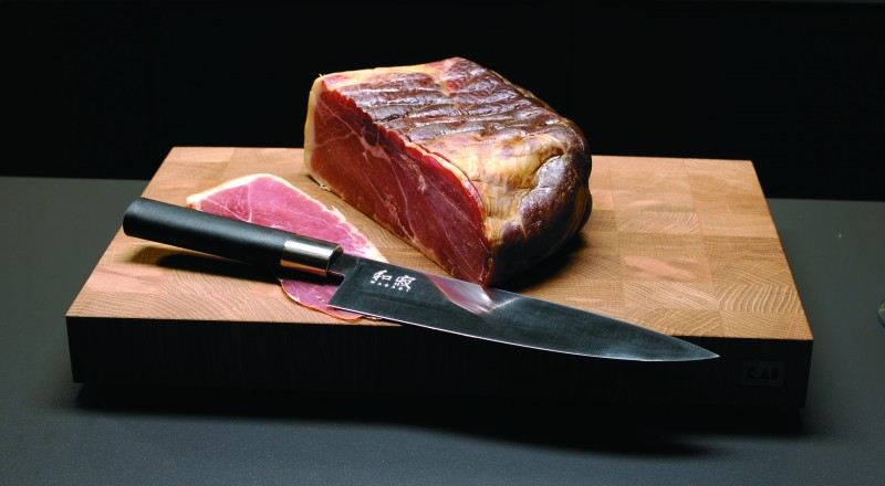 Ein großes Stück gepökeltes Fleisch und eine Scheibe auf einem Holzbrett mit einem Küchenmesser.