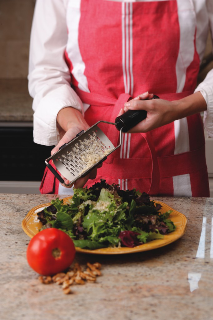 Person in roter Schürze reibt Käse über einen Salat auf einem gelben Teller mit Tomate und Walnüssen.