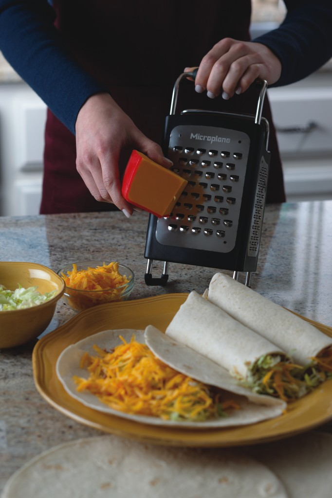 Eine Person reibt Cheddar-Käse für Tacos mit Salat auf einen gelben Teller.