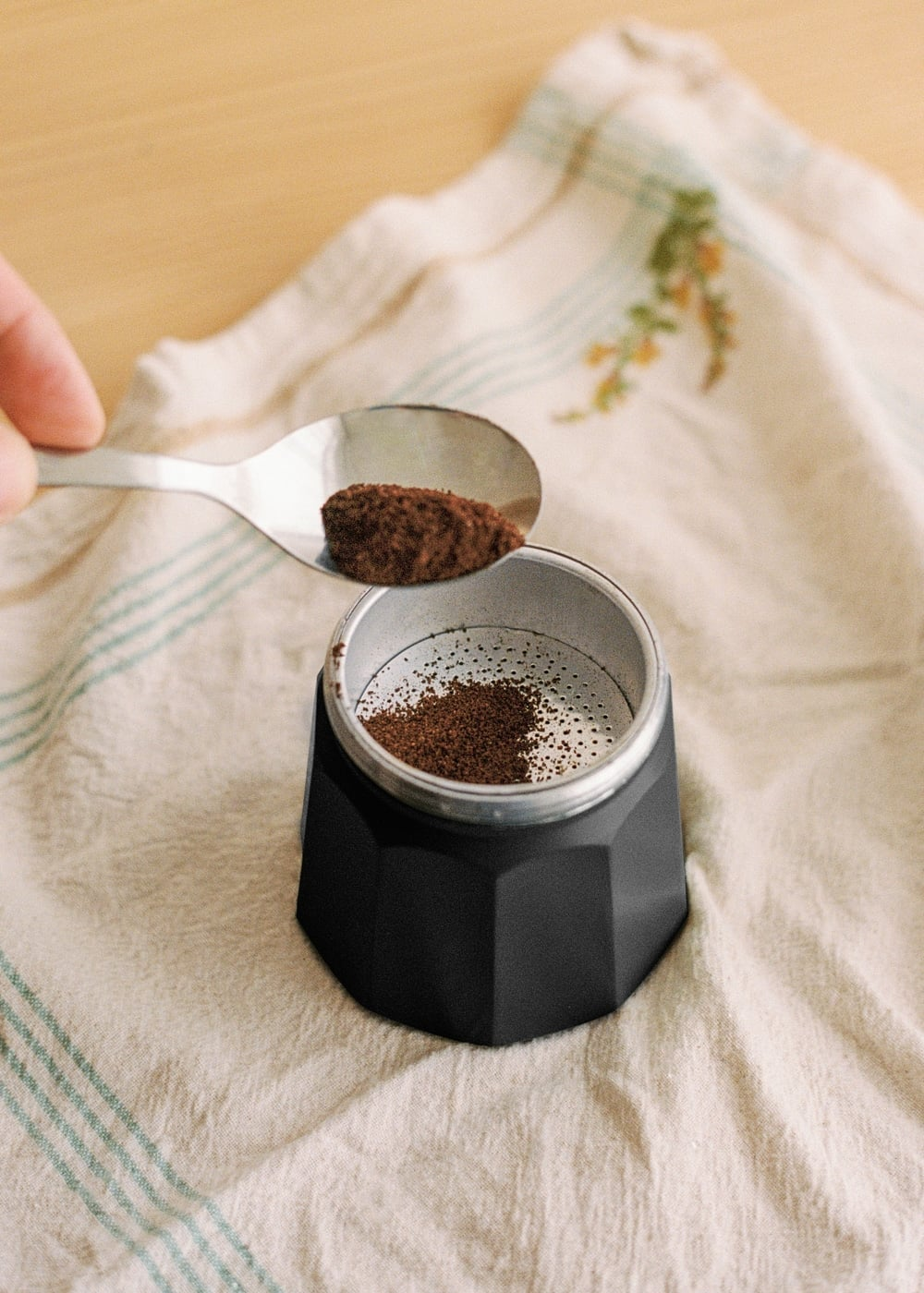 Una cuchara añade café molido al filtro metálico de una cafetera italiana negra sobre un paño.