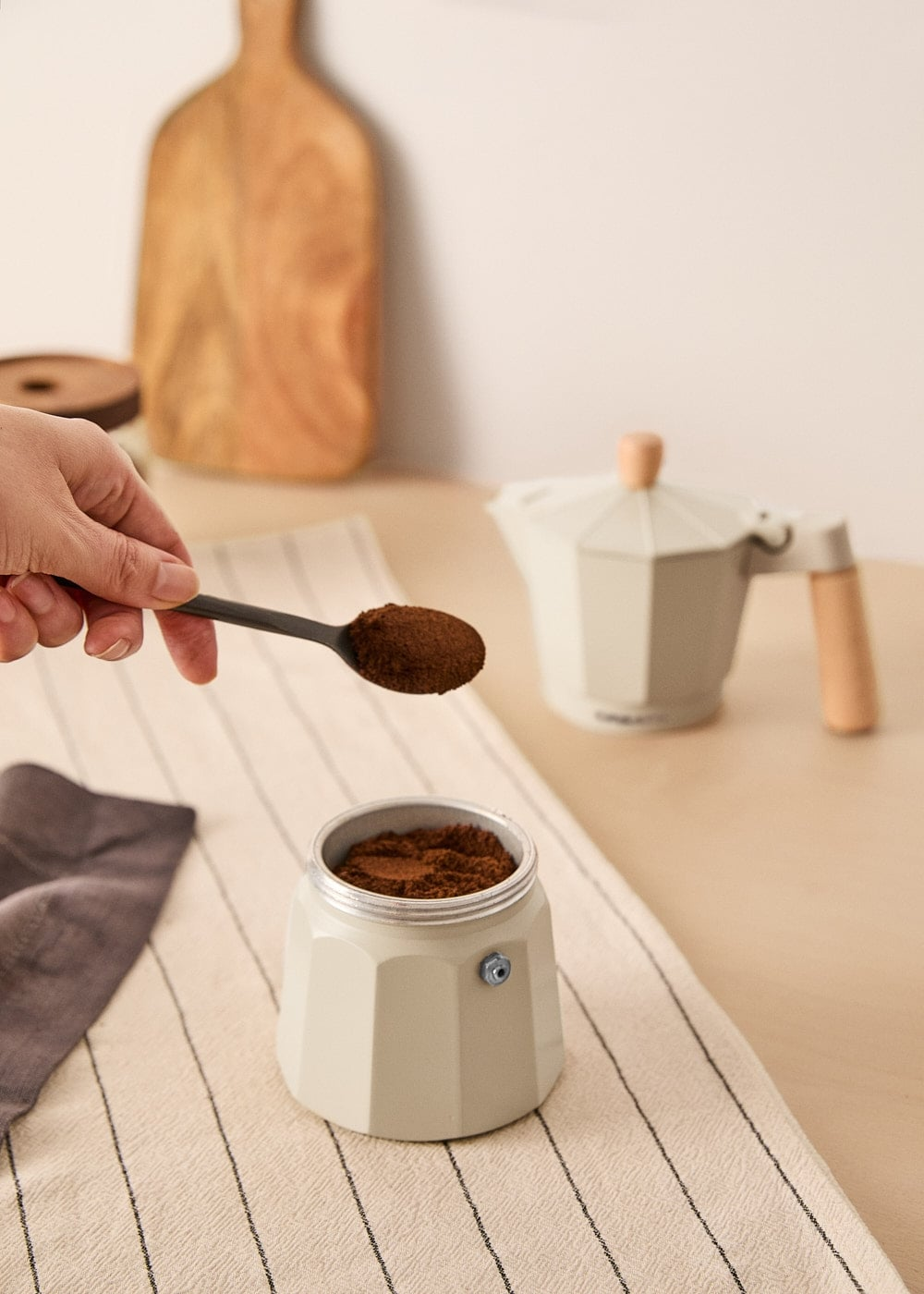 Una mano sostiene una cuchara con café molido sobre el recipiente inferior de una cafetera clara.