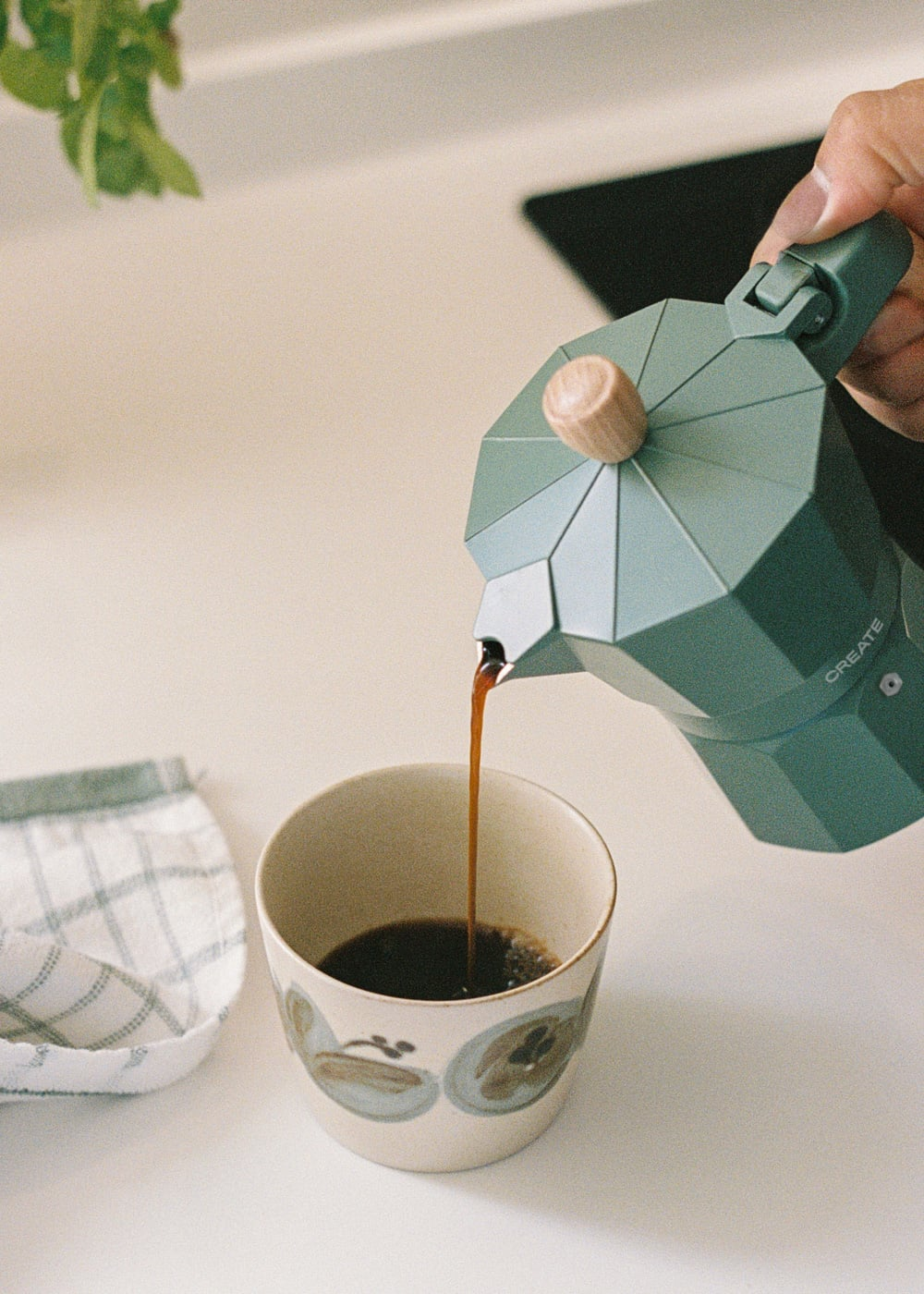Una cafetera italiana verde vierte café en una taza estampada sobre una mesa.