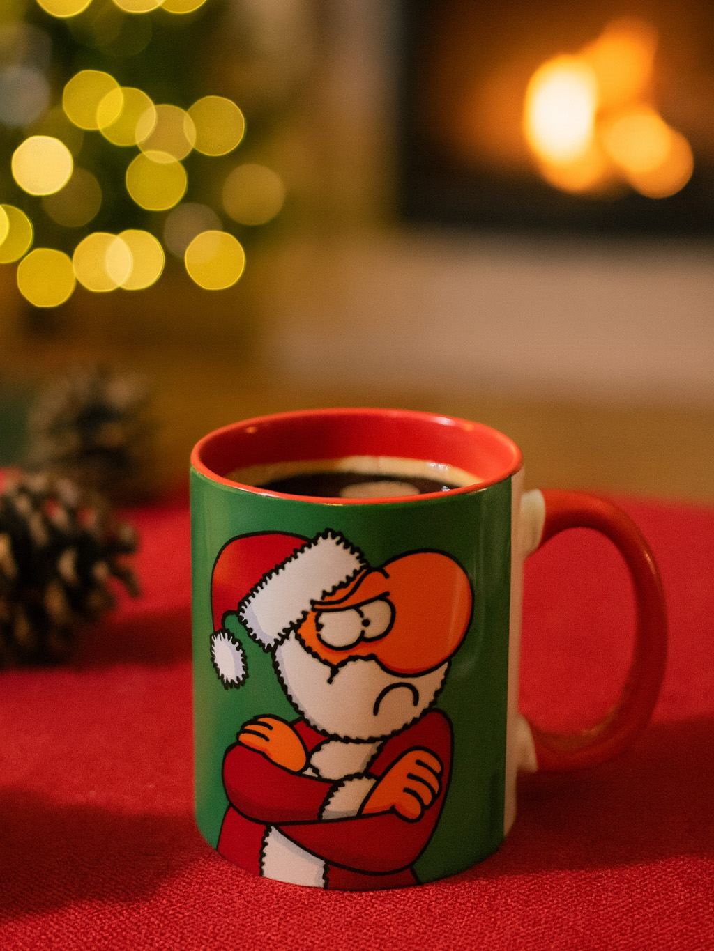 Eine grüne und rote Tasse mit grimmigem Weihnachtsmann auf rotem Tisch vor Kamin und Lichtern.