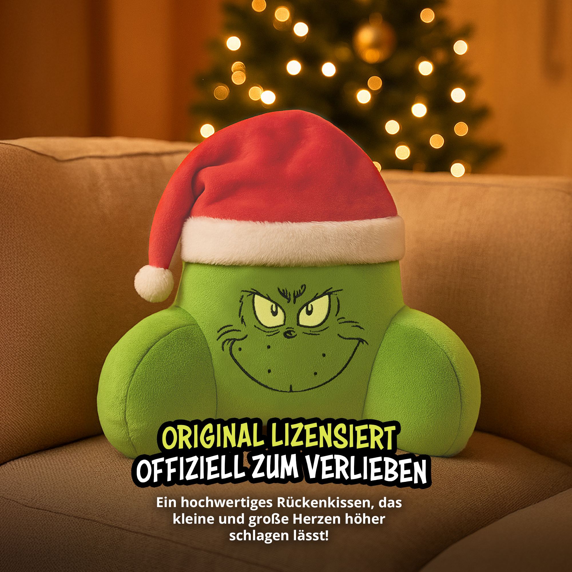 Ein grünes Kissen mit Grinch-Gesicht und Weihnachtsmütze sitzt auf einem Sofa vor einem beleuchteten Weihnachtsbaum.