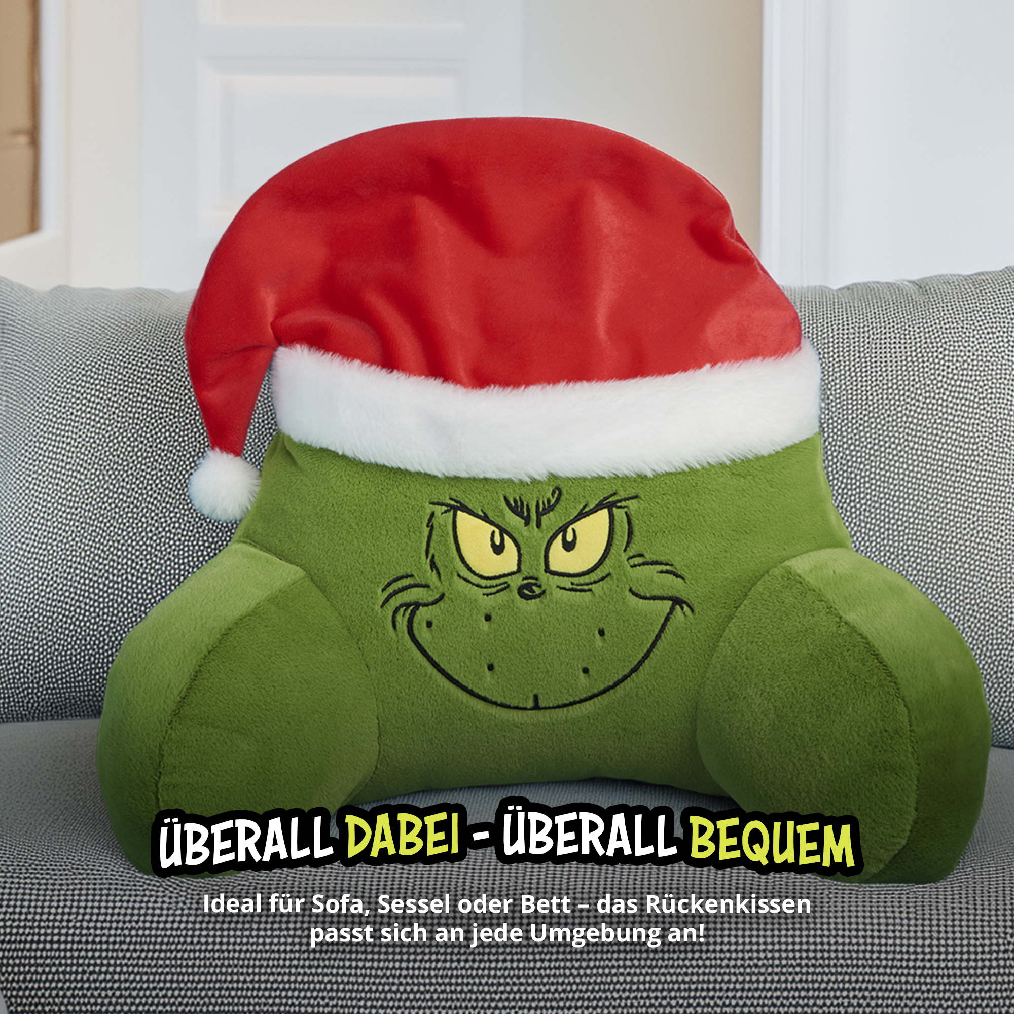 Grünes Grinch Rückenkissen mit rotem Weihnachtsmannhut auf einem grauen Sofa.
