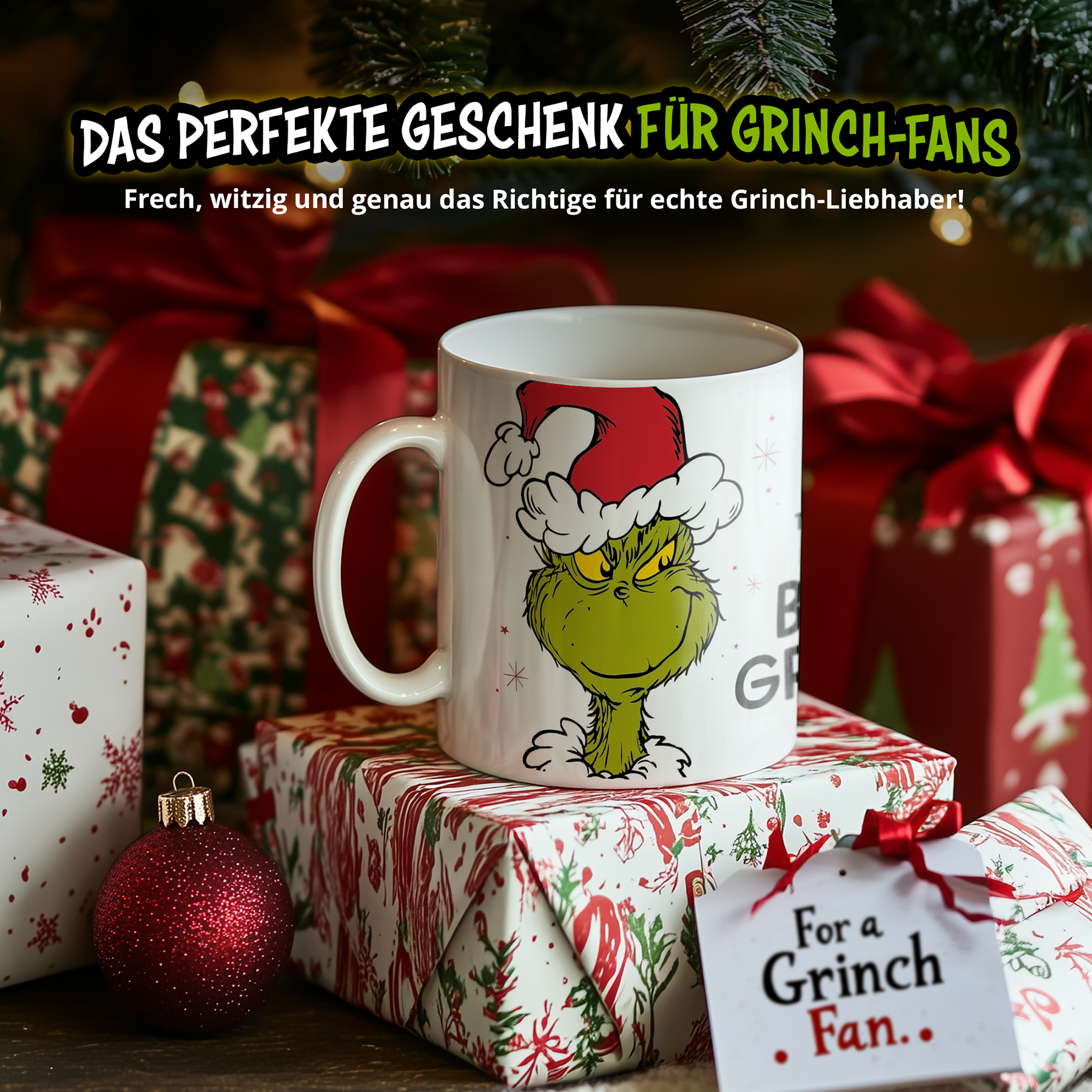 Eine weiße Tasse mit Grinch-Motiv steht auf Weihnachtsgeschenken neben einer roten Kugel vor einem Tannenbaum.