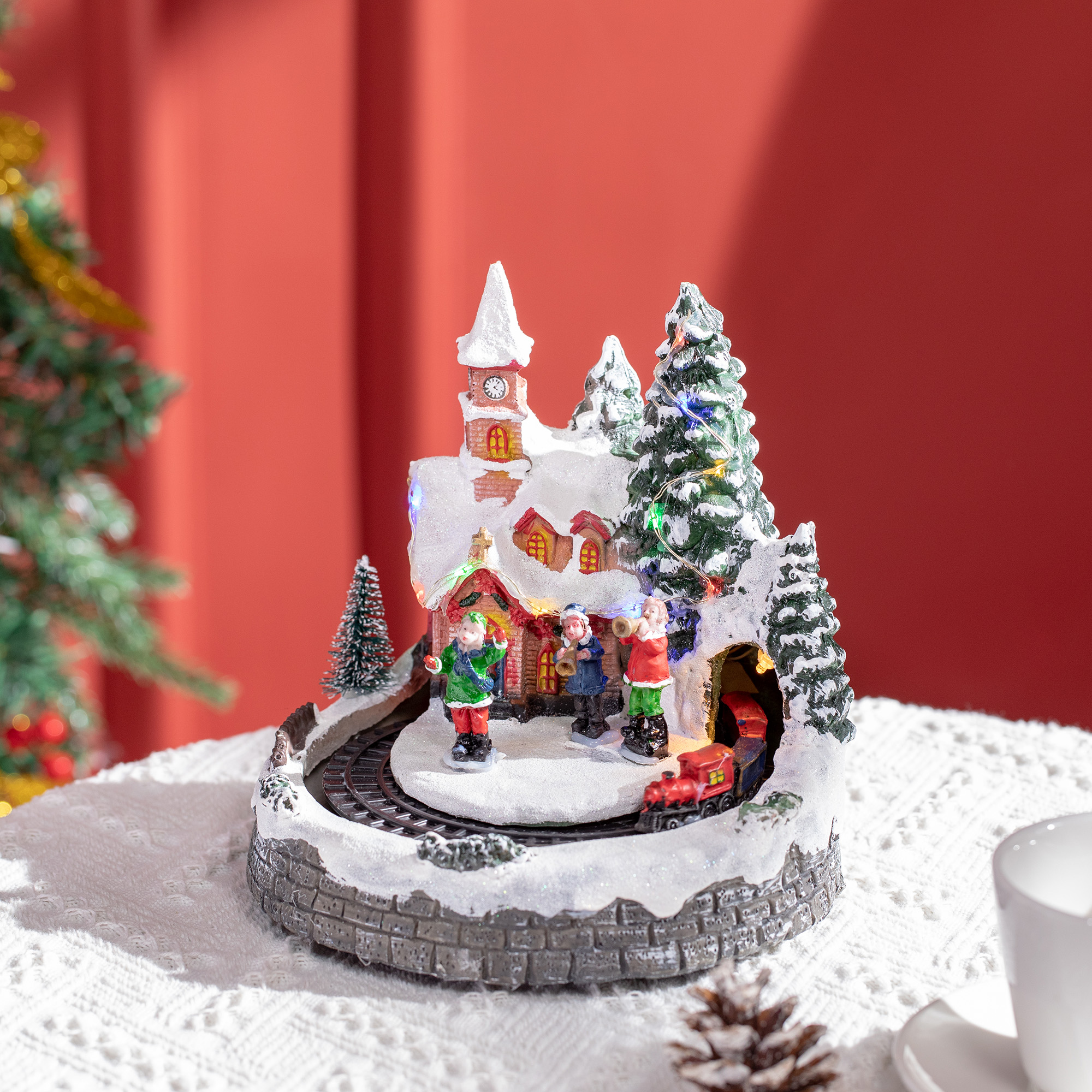 Pueblo navideño cubierto de nieve con figuras, una iglesia y un pequeño tren en una base iluminada.