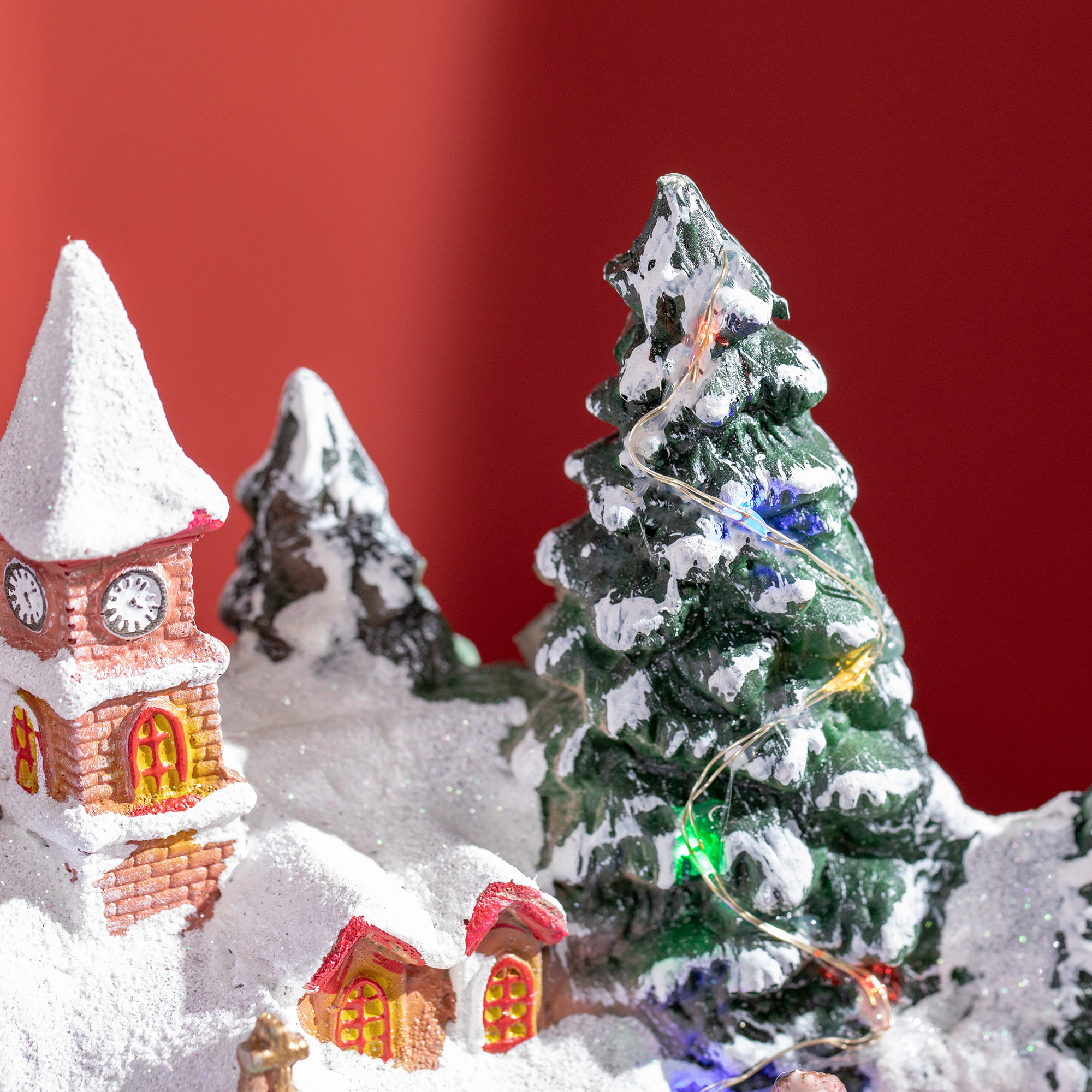 Miniatura de pueblo invernal nevado con una iglesia, un árbol de Navidad y luces sobre fondo rojo.