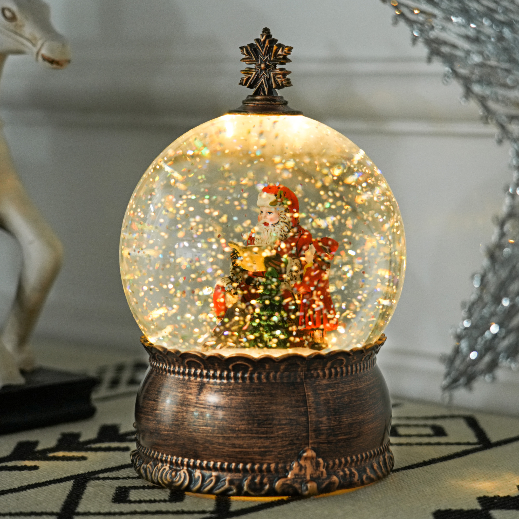 Una bola de nieve iluminada con base ornamentada muestra a Santa Claus con regalos dorados y nieve brillante.