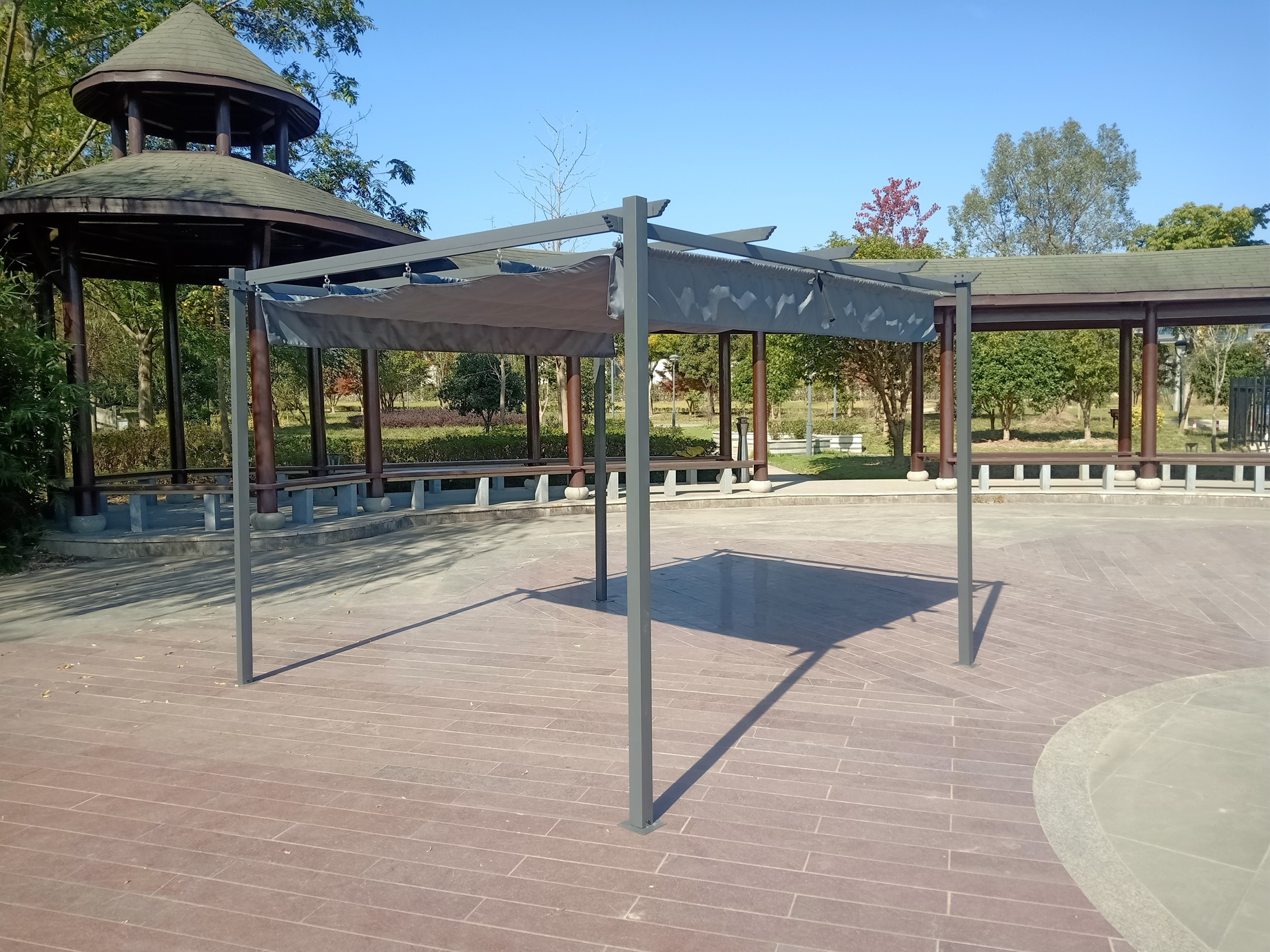 Nowoczesna szara metalowa pergola z daszkiem stoi na wybrukowanym placu z altanami i drzewami w tle.