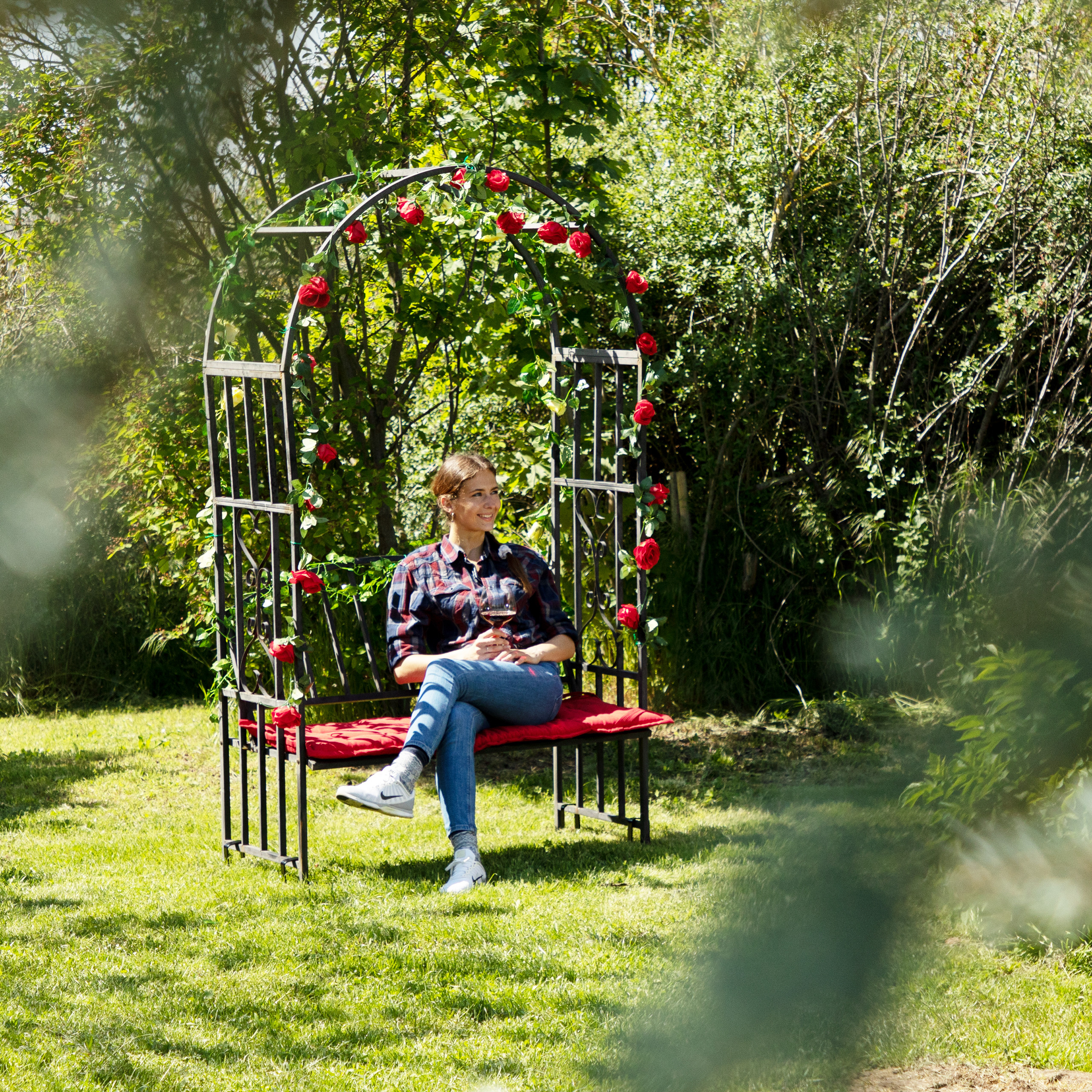 Eine Frau sitzt auf einer Gartenbank unter einem Rosenbogen und hält ein Glas Wein in der Hand.