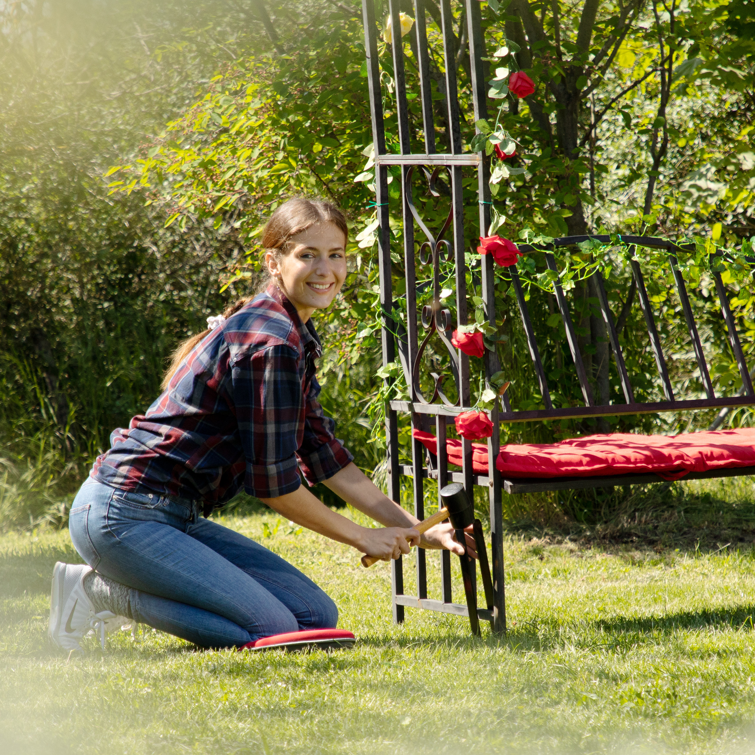 Eine Frau in kariertem Hemd kniet im Gras und hämmert an einer roten Rosen-Gartenbank.
