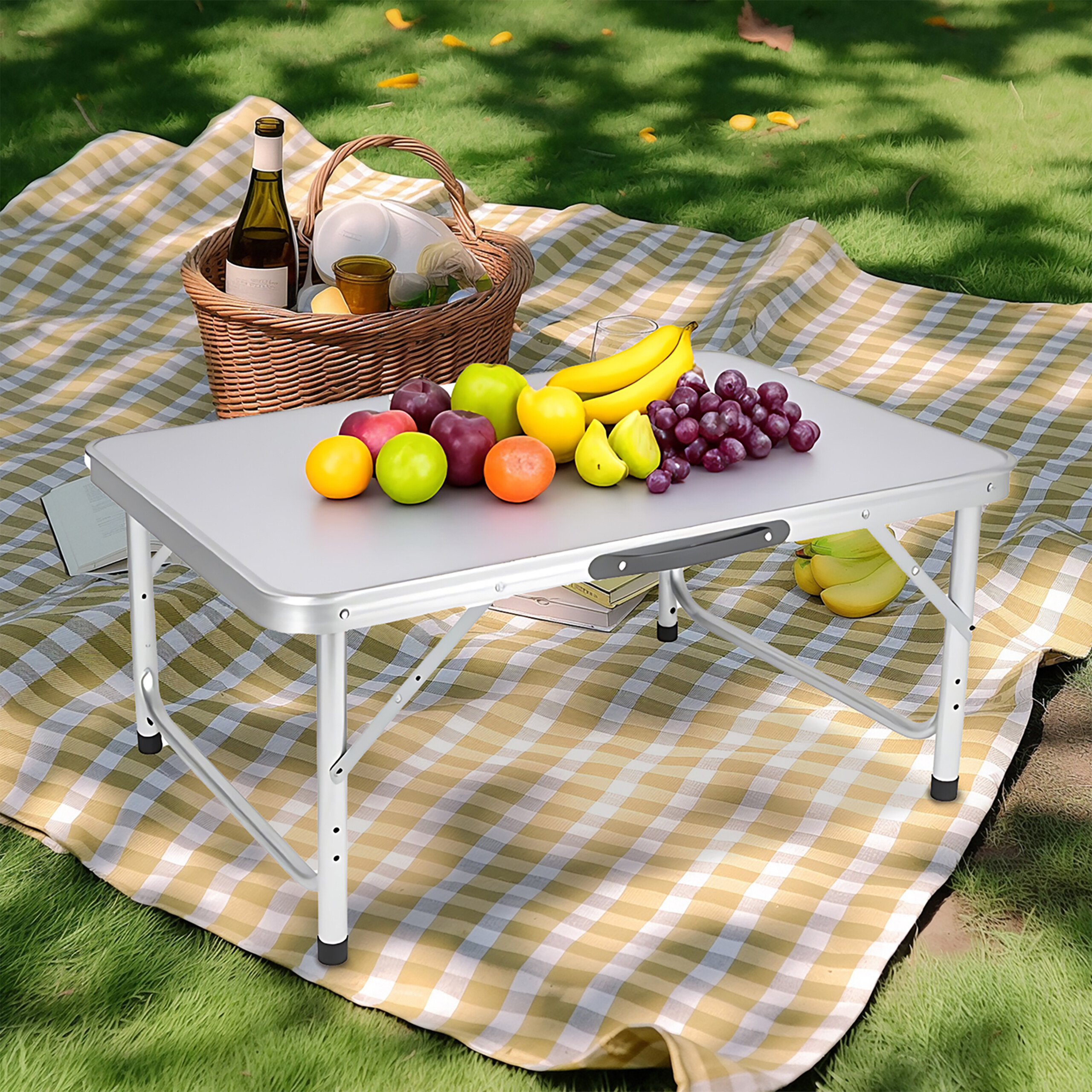Ein kleines Tischchen mit Obst steht auf einer karierten Picknickdecke im Gras, dahinter ein Korb mit Flasche.