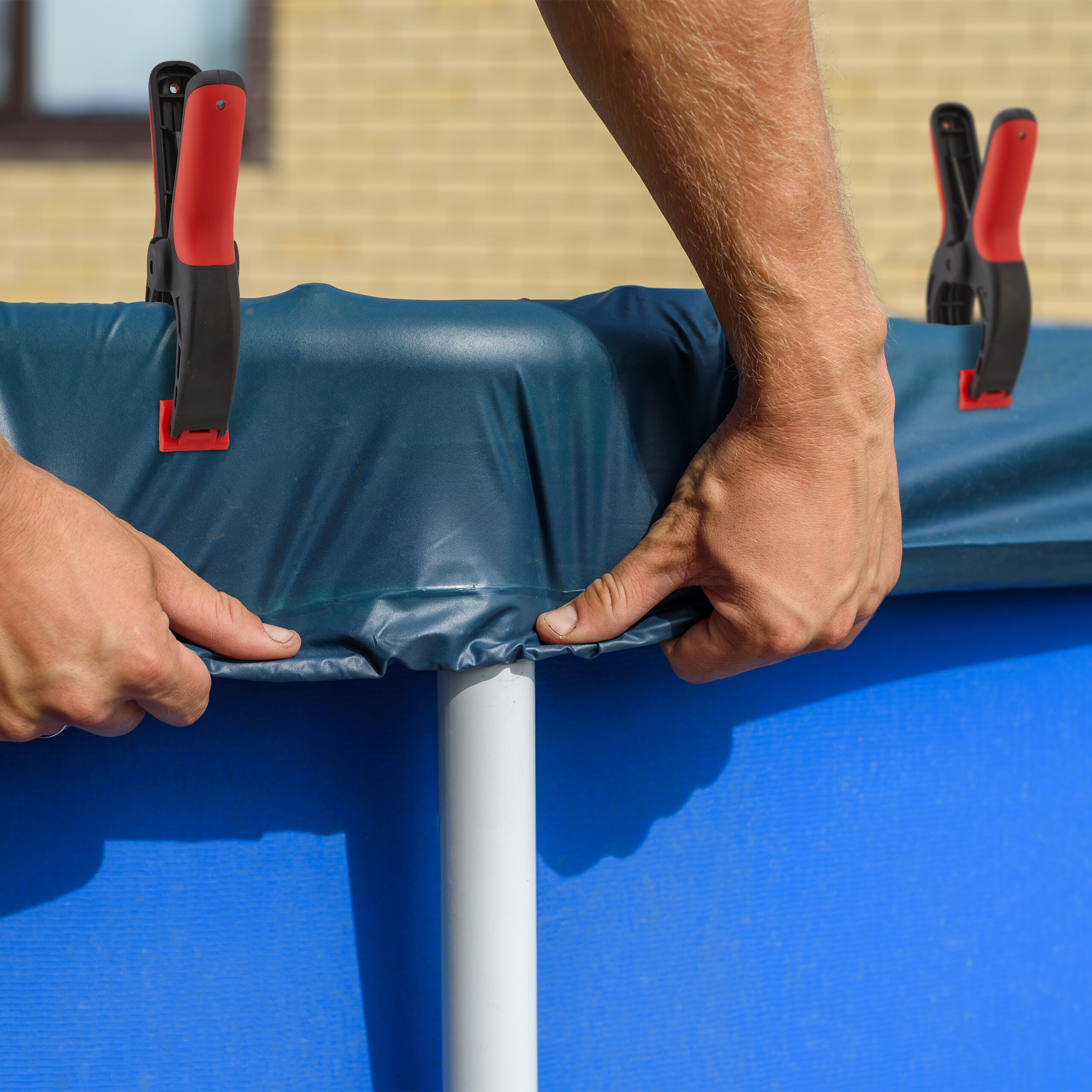 Person sichert eine blaue Poolabdeckung mit roten und schwarzen Klemmen.