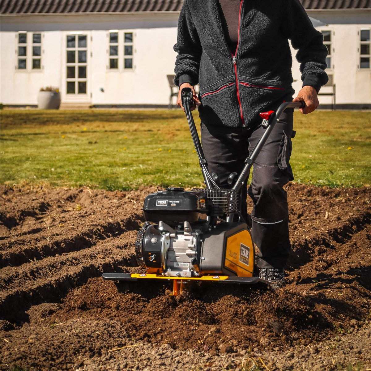 Eine Person in schwarzer Kleidung bedient eine Gartenfräse auf einem erdbedeckten Beet vor einem weißen Haus.