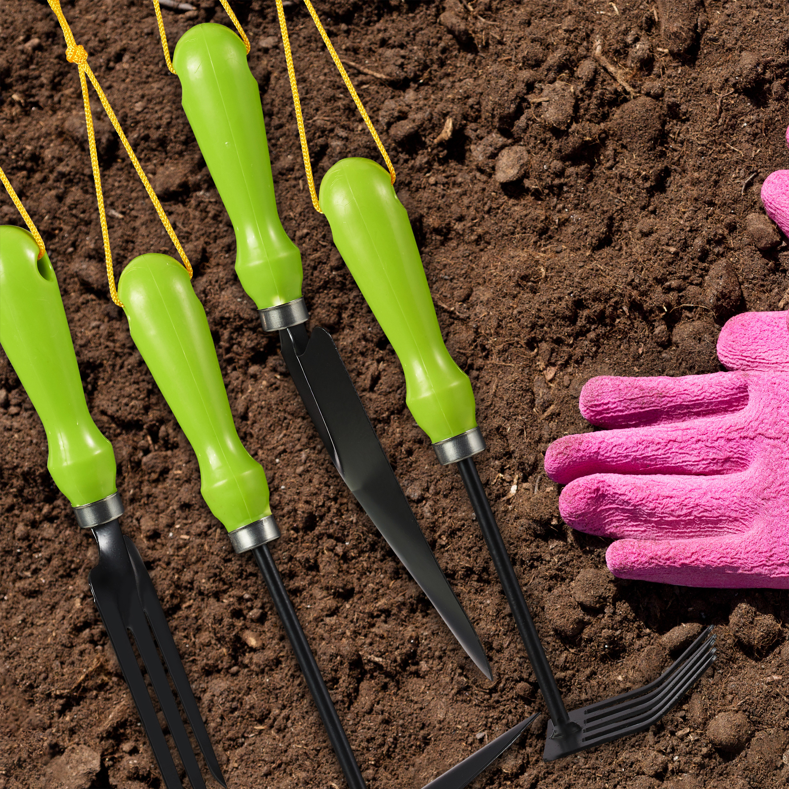 Vier Gartengeräte mit grünen Griffen und eine pinke Handschuh auf brauner Erde.