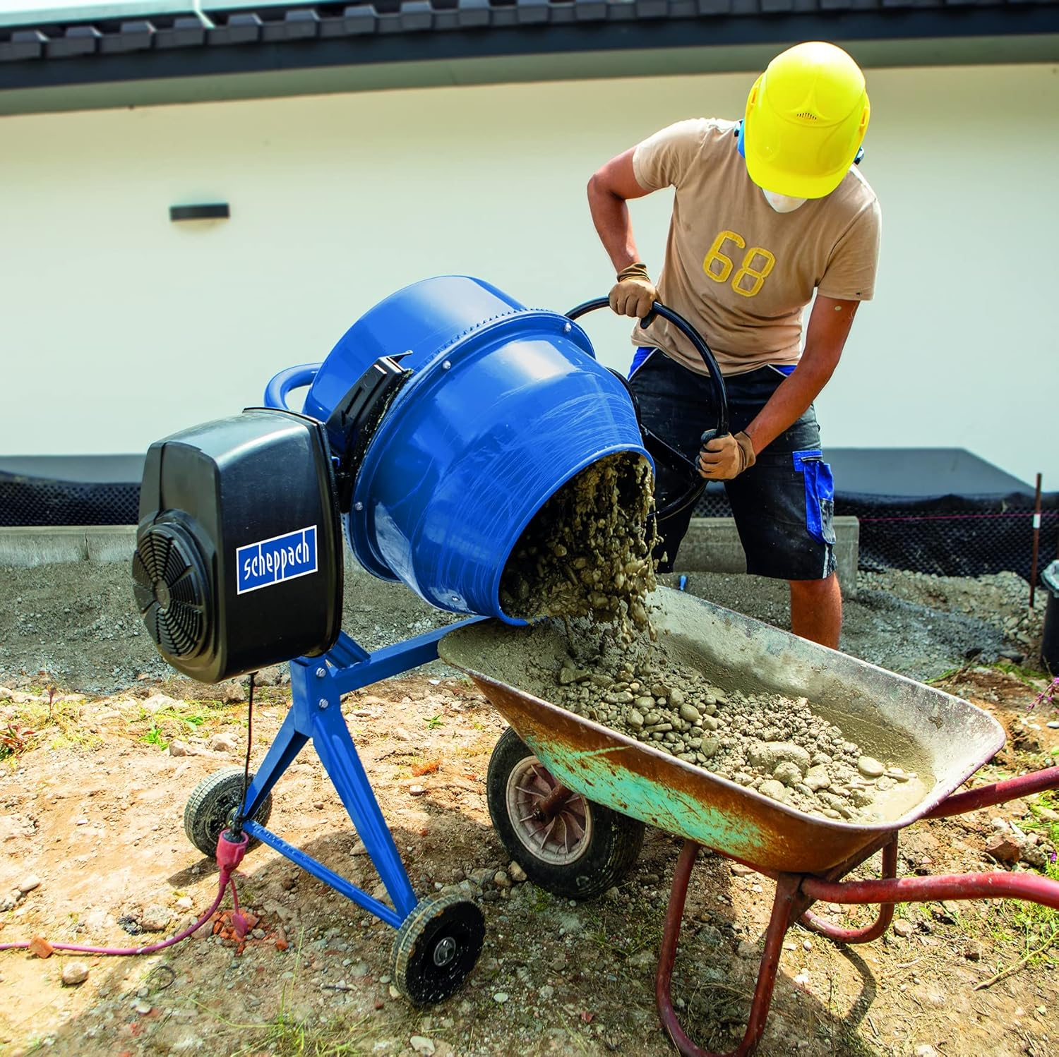 Ein Arbeiter in gelbem Helm kippt Beton aus einem blauen Mischer in eine Schubkarre auf einer Baustelle.