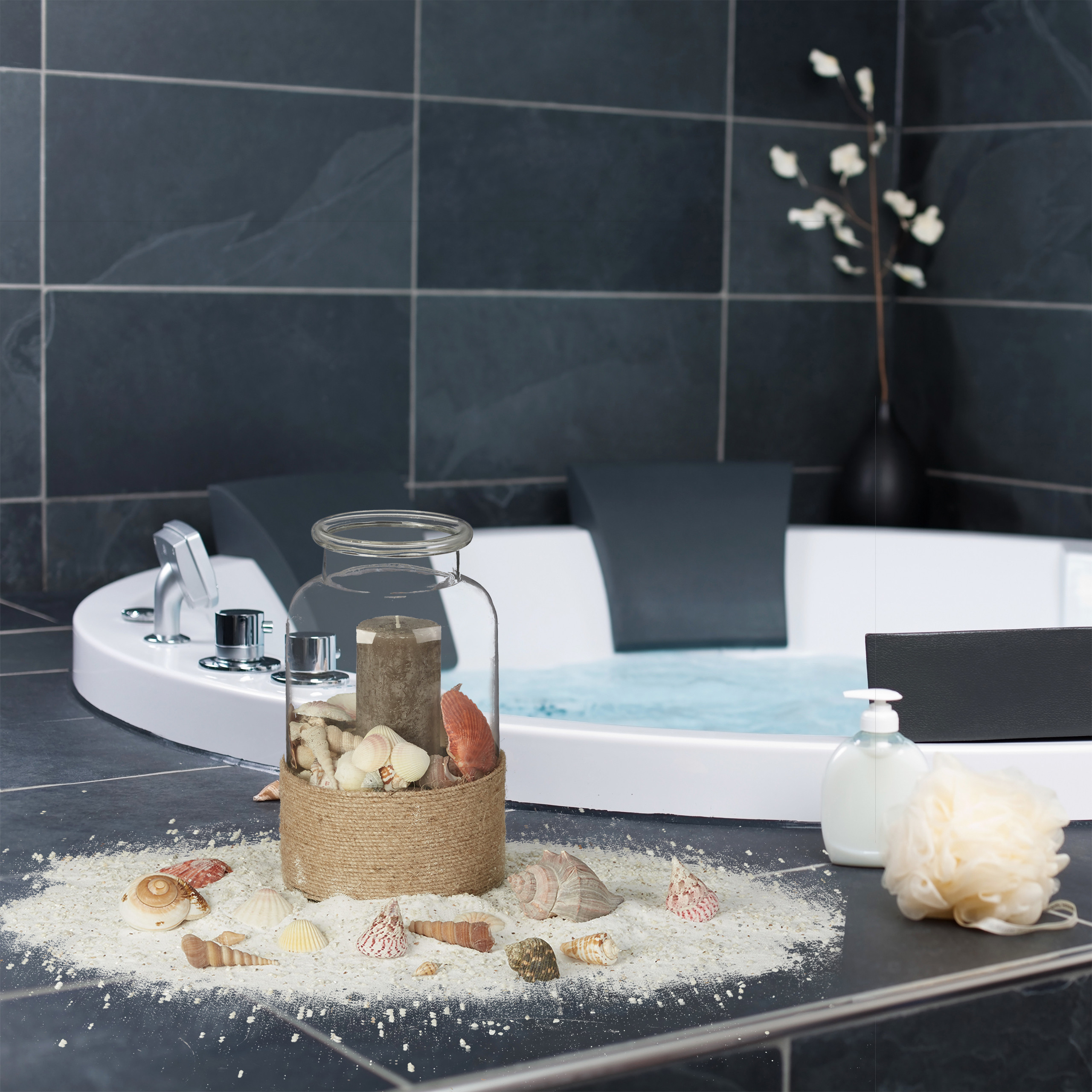 Glasgefäß mit Kerze und Muscheln auf Sand vor einer runden Badewanne mit dunkelgrauen Fliesen.