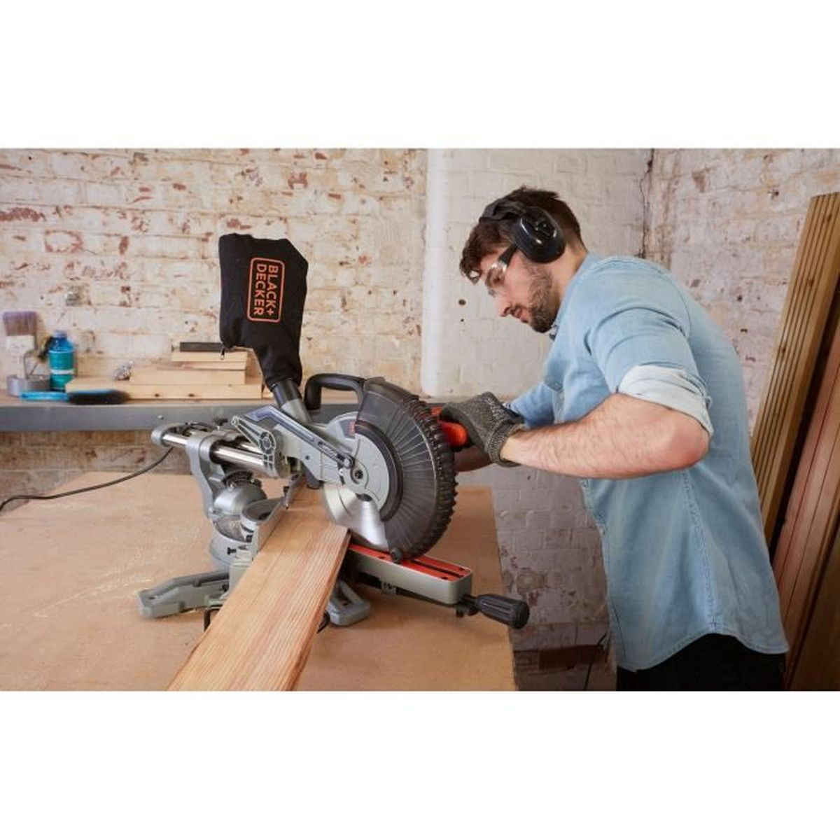Ein Mann mit Gehörschutz und Schutzbrille schneidet ein Holzbrett mit einer schwarzen Kappsäge in einer Werkstatt.