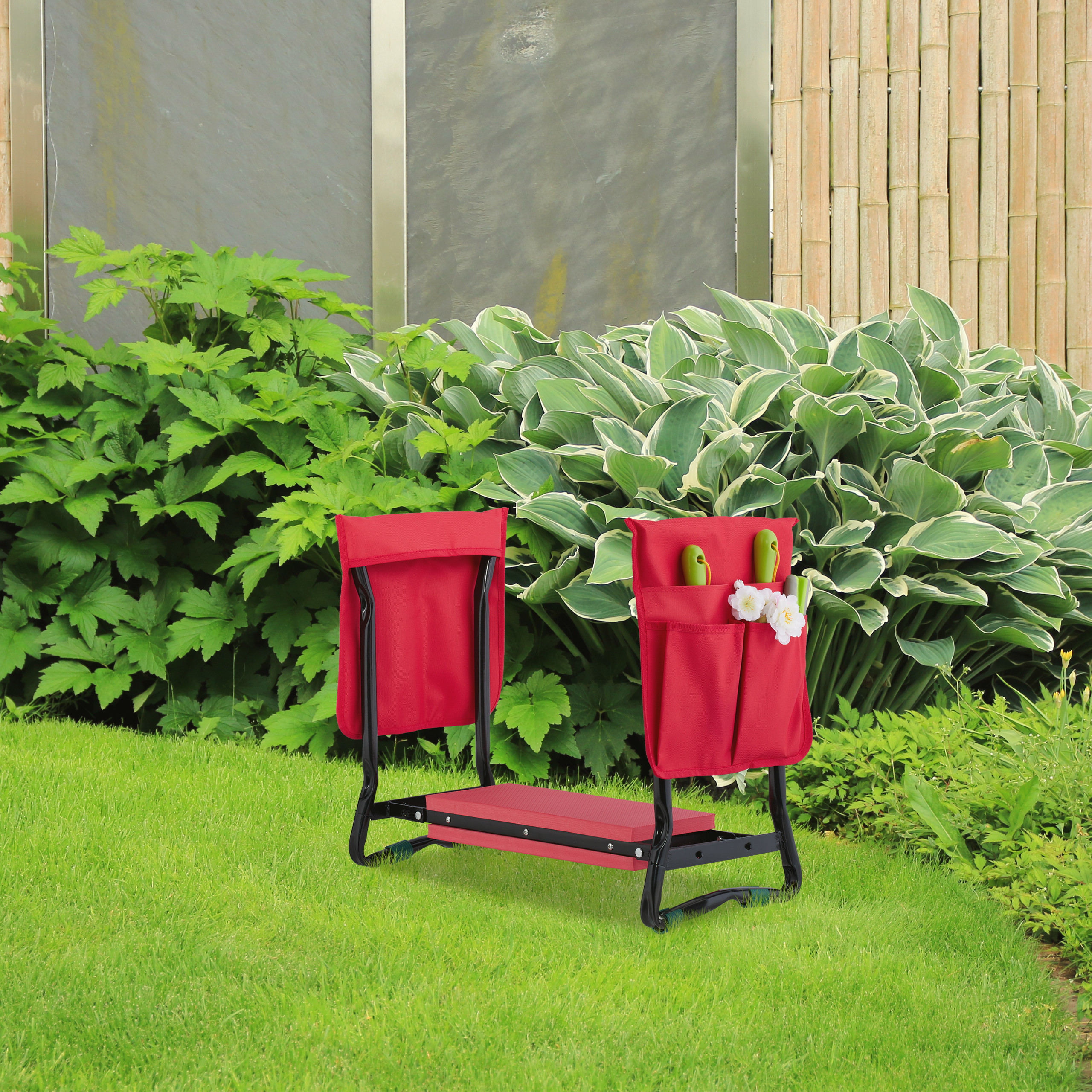 Ein roter Gartenhocker und Knieschoner mit Werkzeugtasche steht auf grünem Rasen vor üppigen Pflanzen.