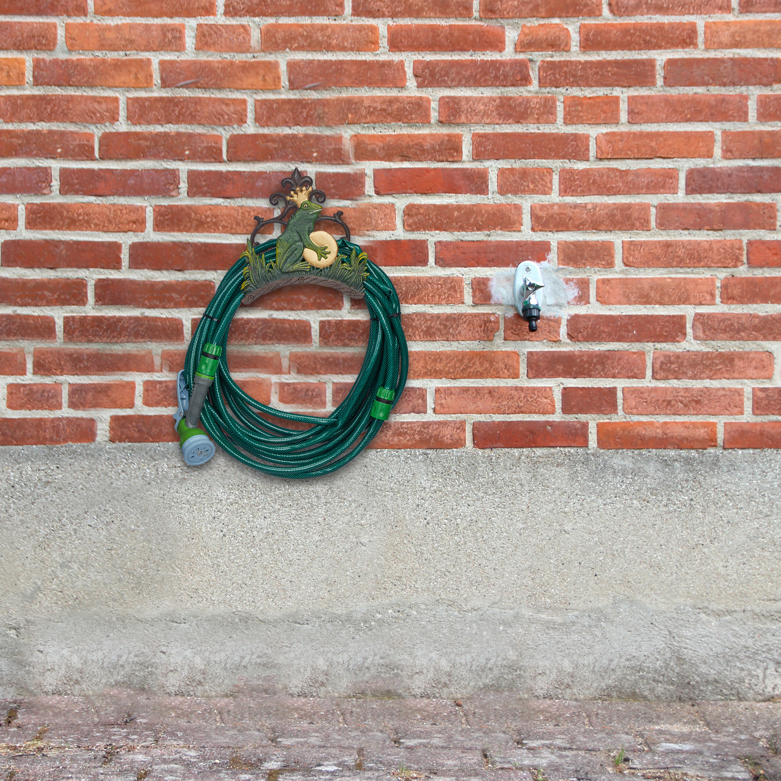 Ein aufgerollter grüner Gartenschlauch hängt an einer froschverzierten Halterung neben einem Wasserhahn an einer Backsteinwand.