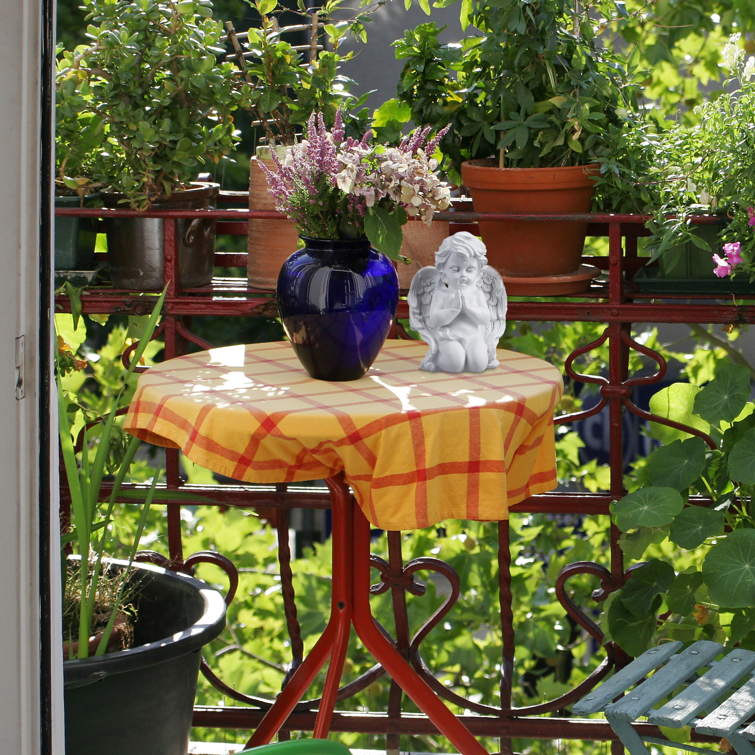 Ein Balkon mit einem runden Tisch mit gelb-roter Decke, einer blauen Vase und einer Engelsfigur vor Grünpflanzen.