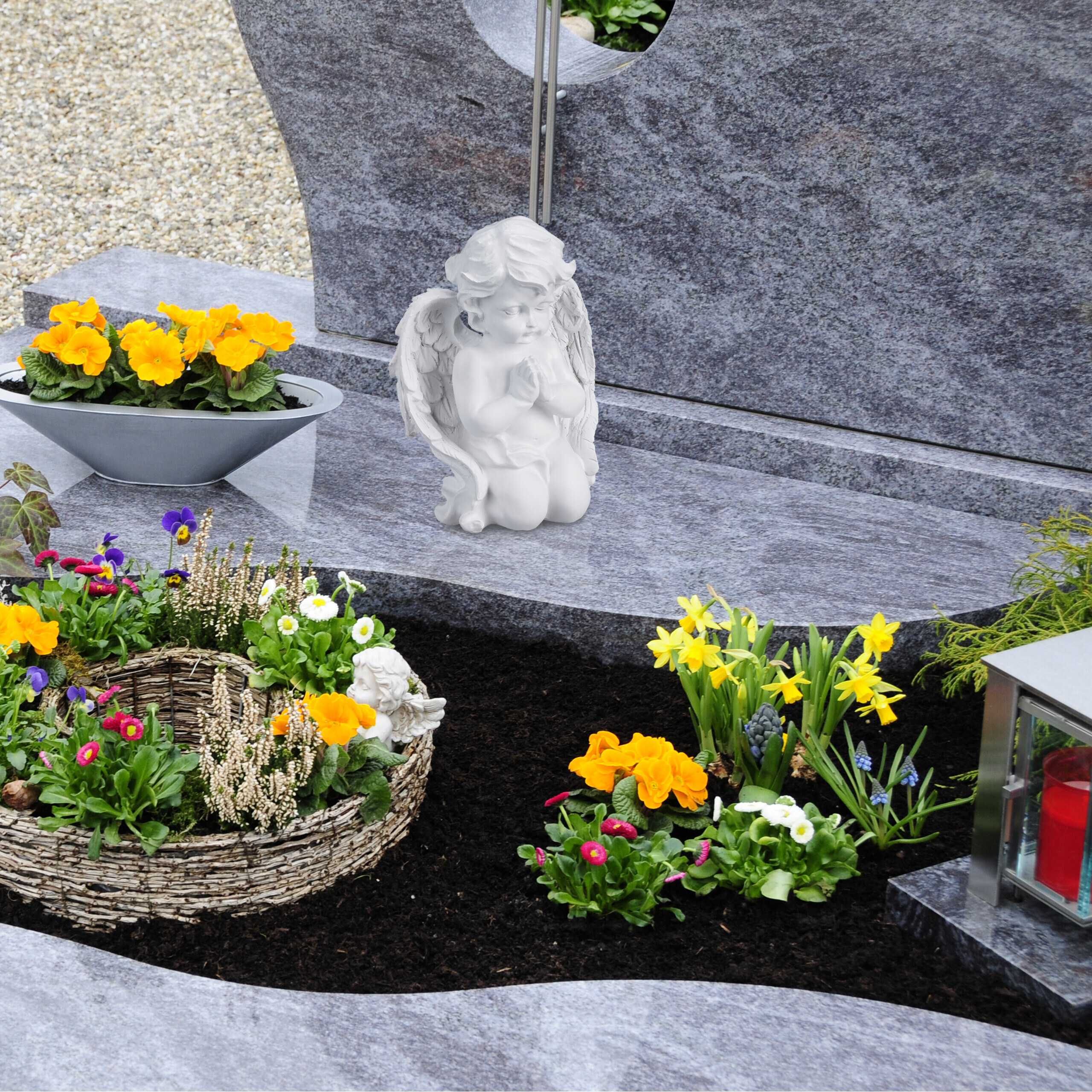 Ein Grabstein mit einem betenden Engel, gelben Blumen in einer Schale, bunten Pflanzen und einer roten Grabkerze.