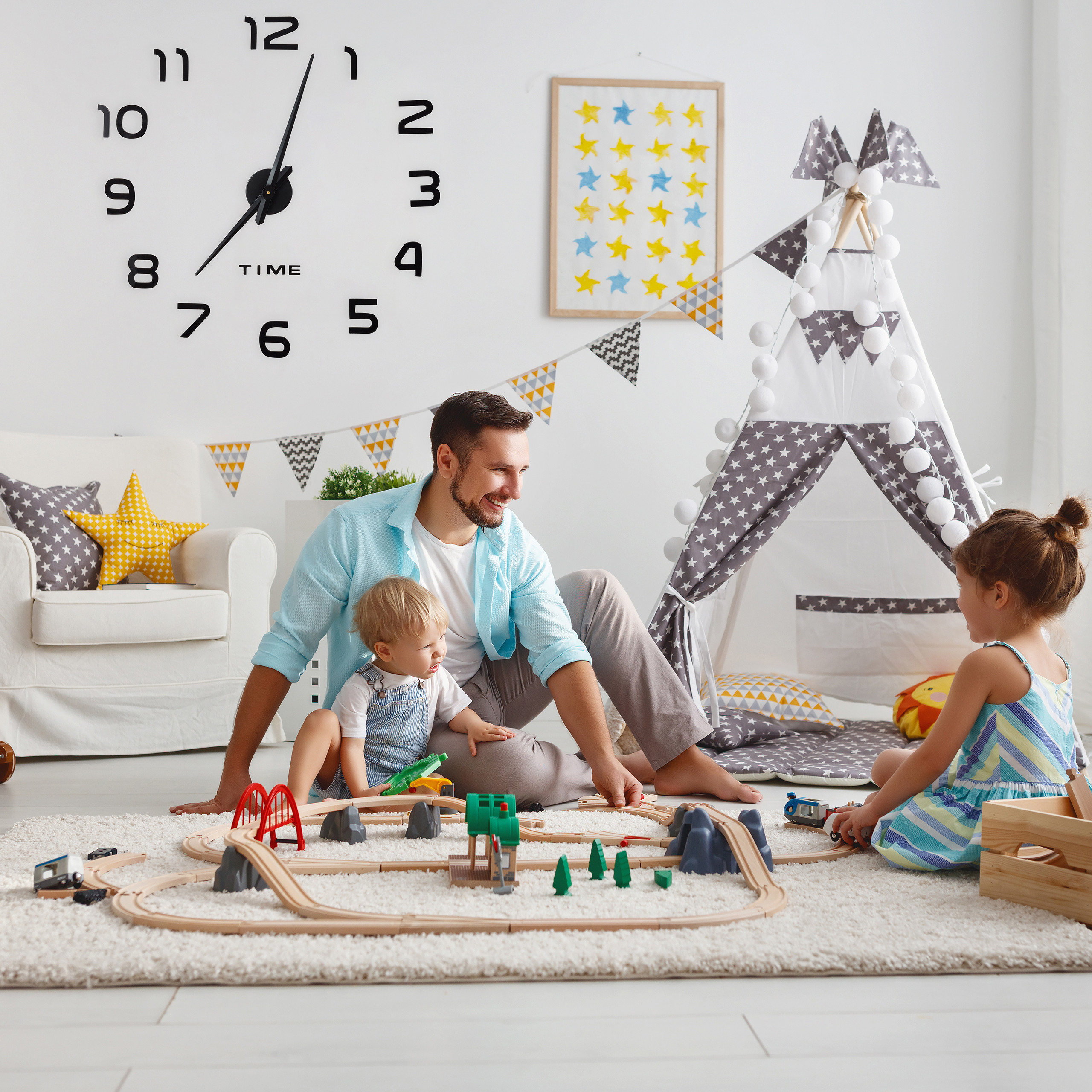 Vater und zwei Kinder spielen im hellen Zimmer mit einer Holzeisenbahn. An der Wand hängt eine große Uhr.