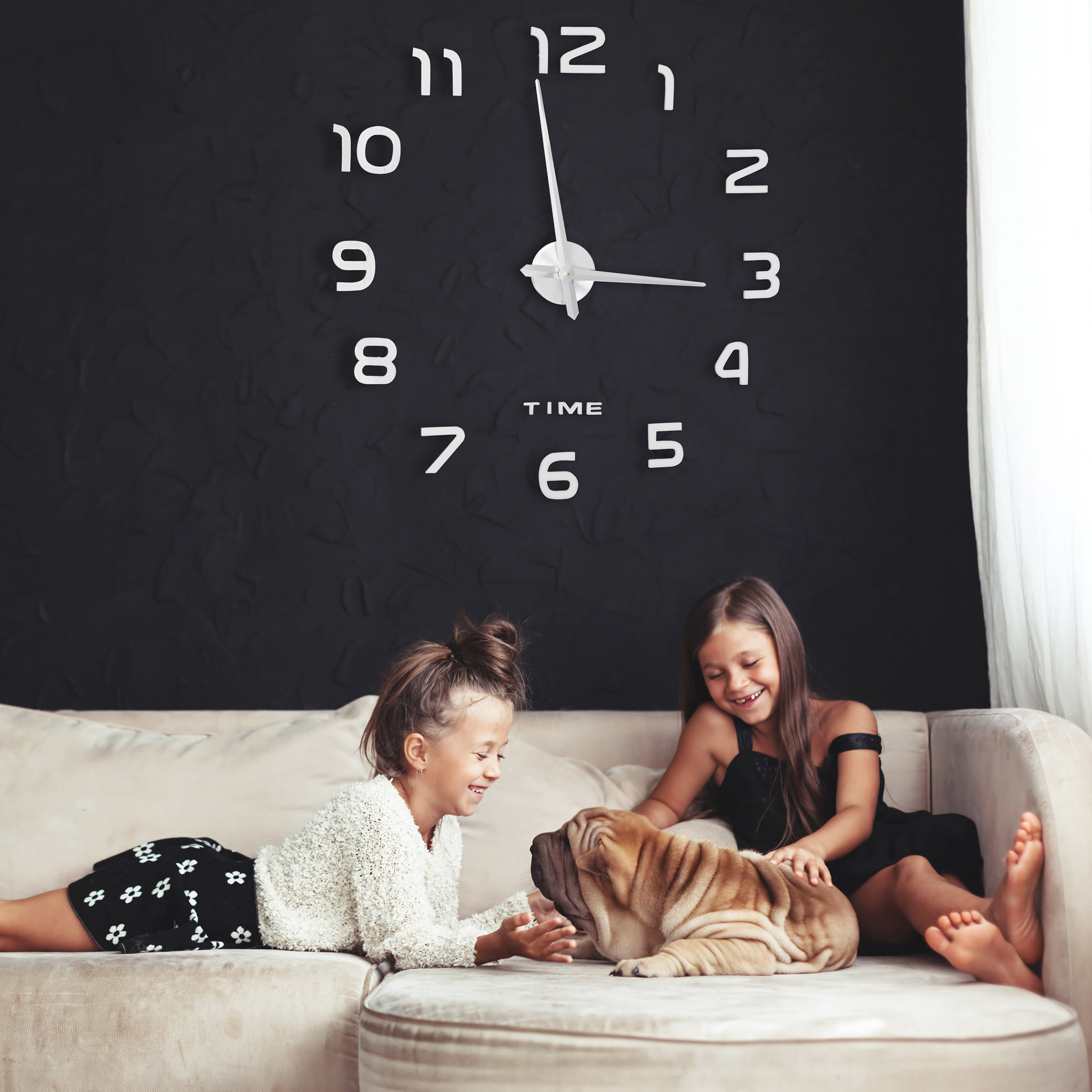 Zwei Mädchen lächeln, während sie mit einem Shar-Pei-Welpen auf einer hellen Couch unter einer großen Wanduhr spielen.