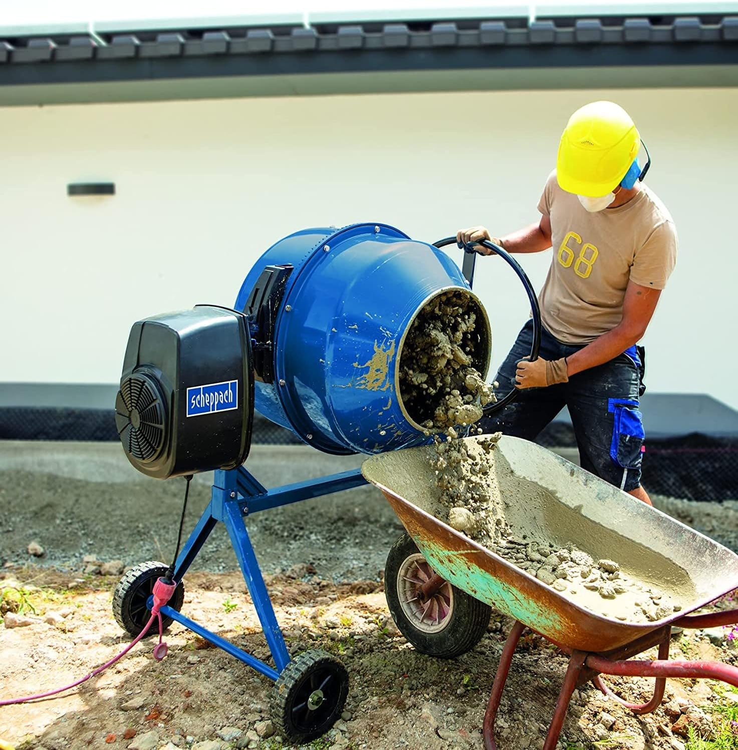 Ein Arbeiter in gelbem Helm gießt Beton aus einer blauen Trommel in eine Schubkarre.