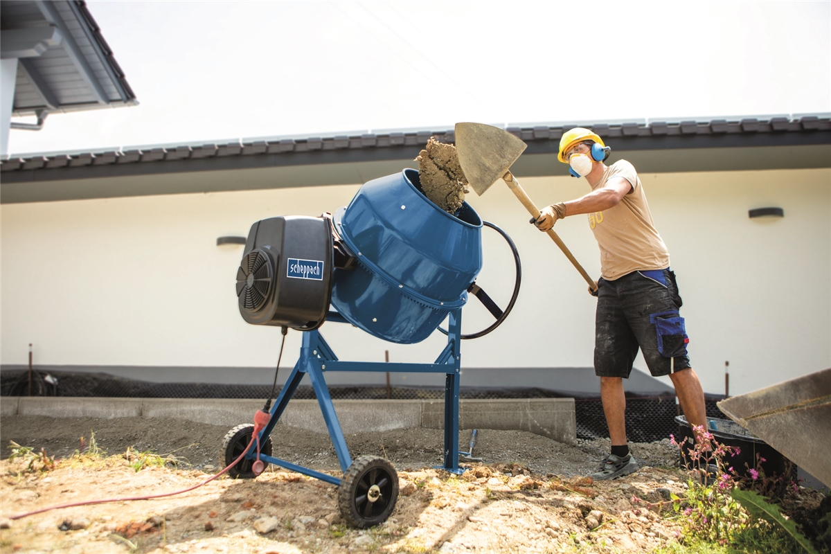 Ein Arbeiter mit Helm füllt Zement in eine blaue Betonmischmaschine auf einer Baustelle.