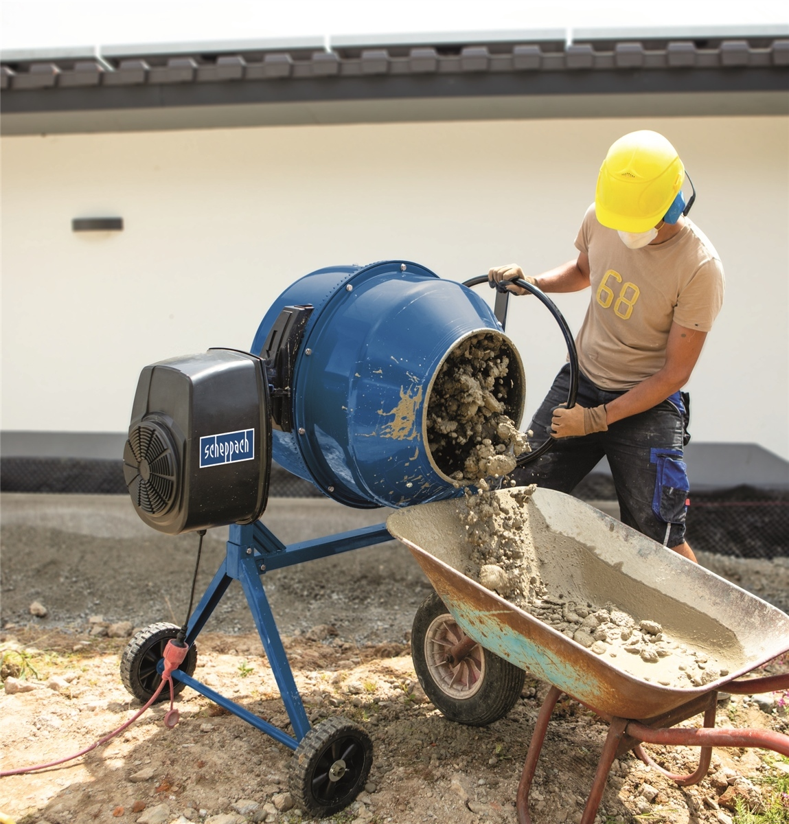 Ein Arbeiter mit gelbem Schutzhelm gießt Beton aus einem blauen Mischer in eine Schubkarre.