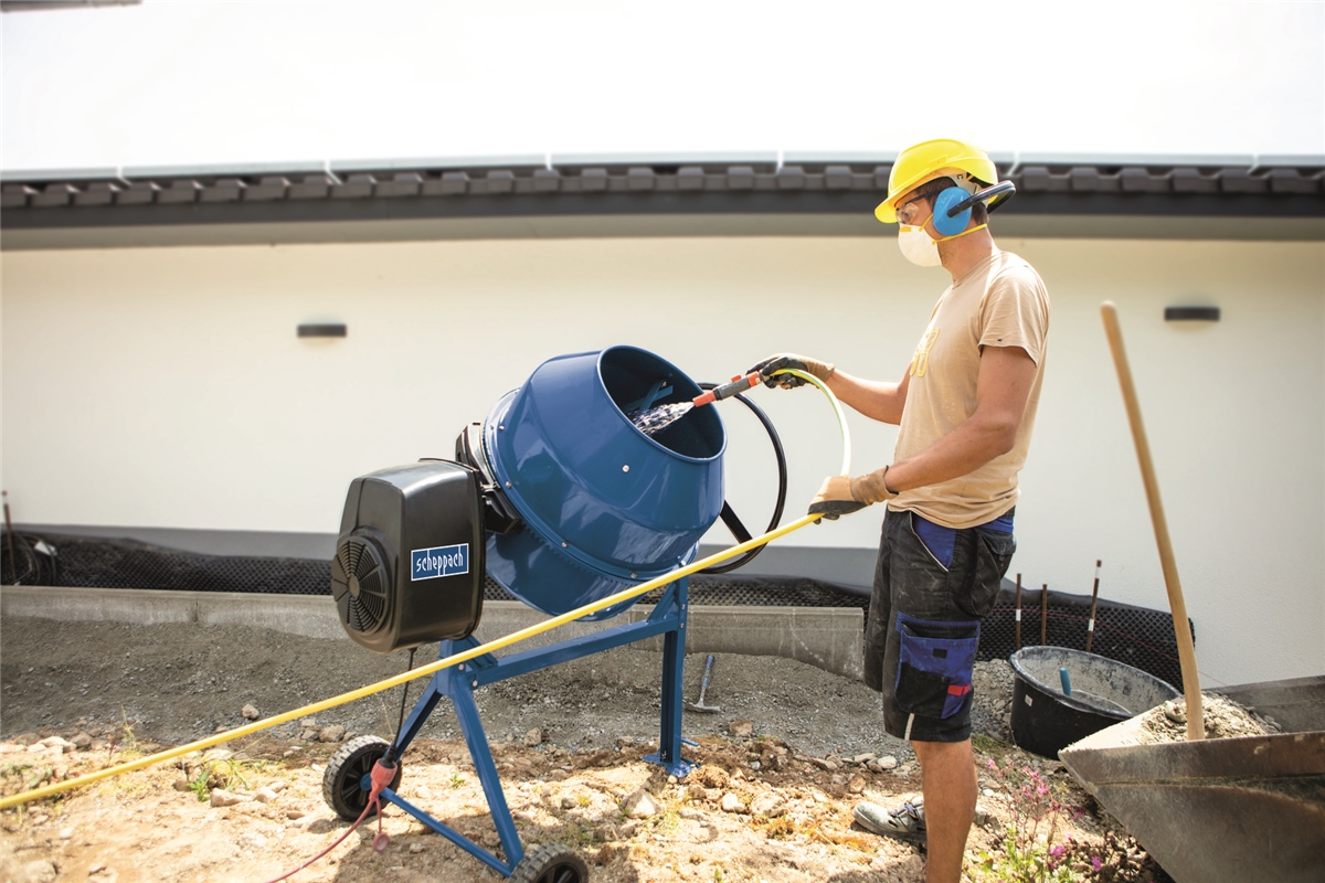 Ein Arbeiter in gelbem Helm füllt Wasser in eine blaue Betonmischmaschine auf einer Baustelle.