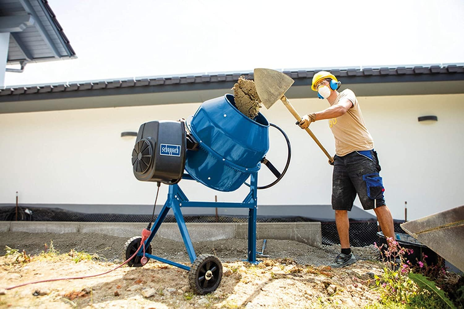 Ein Arbeiter füllt mit einer Schaufel Erde in einen blauen Betonmischer auf einer Baustelle.
