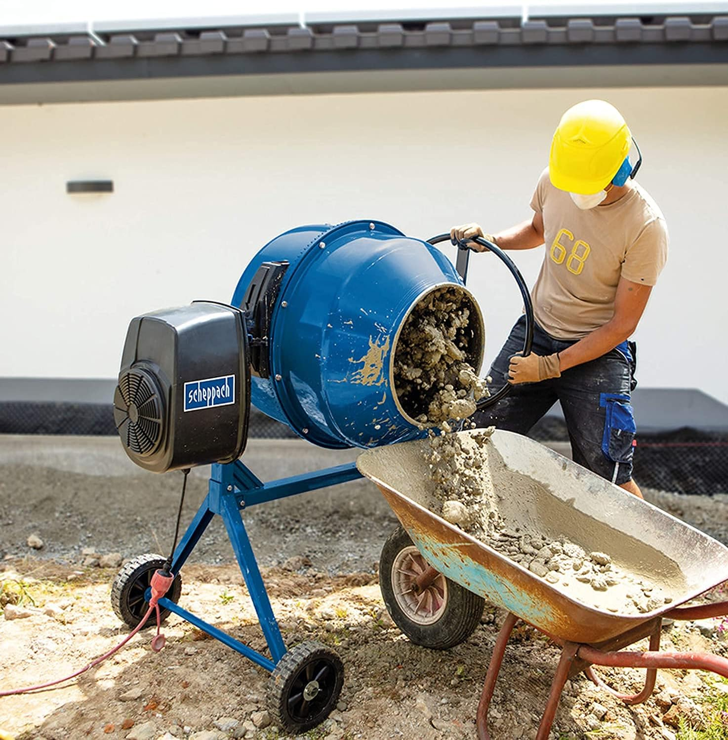 Ein Arbeiter mit Helm gießt Beton aus einer blauen Trommelmischmaschine in eine Schubkarre.
