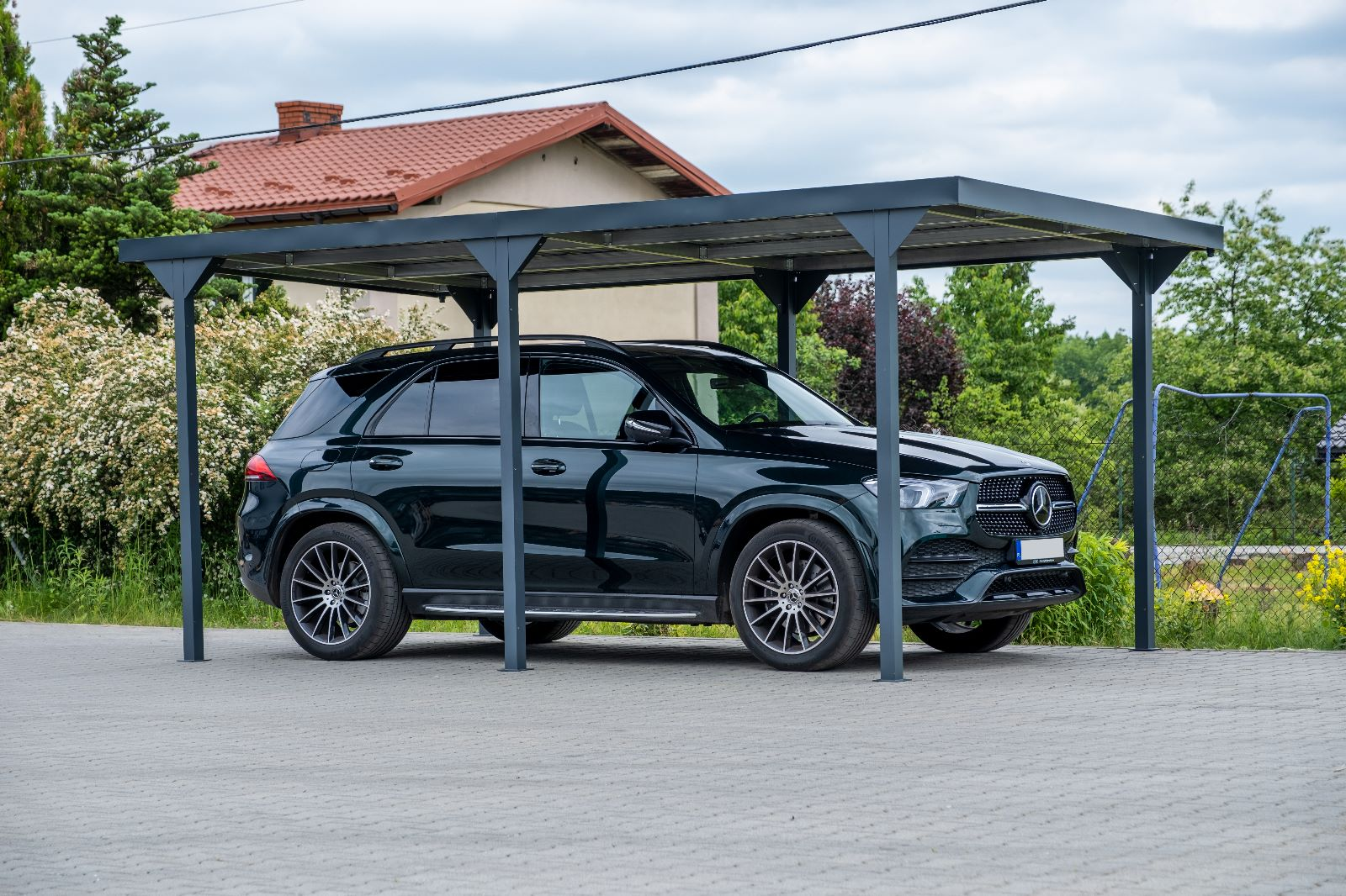 Czarny SUV parkuje pod nowoczesną szarą wiatą na utwardzonym podwórku przed domem.