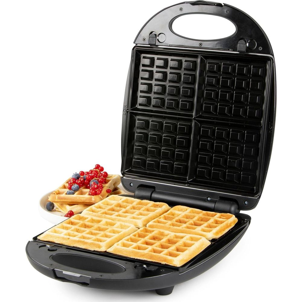 Una piastra per waffle nera aperta cuoce quattro waffle dorati, con bacche fresche accanto.