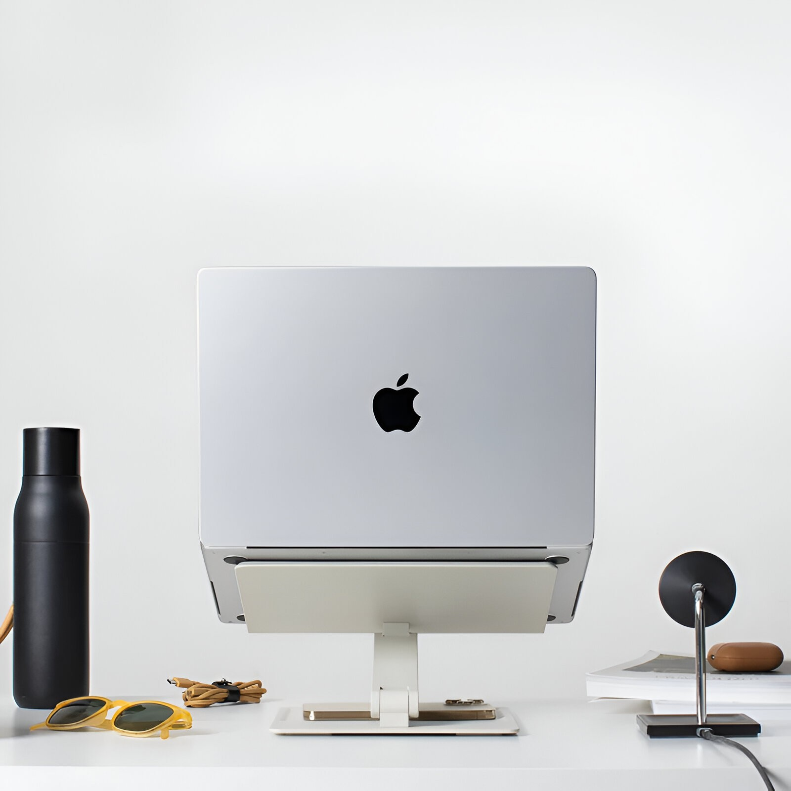 Ein geschlossenes MacBook auf einem weißen Ständer auf einem Schreibtisch, flankiert von einer Flasche und Zubehör.