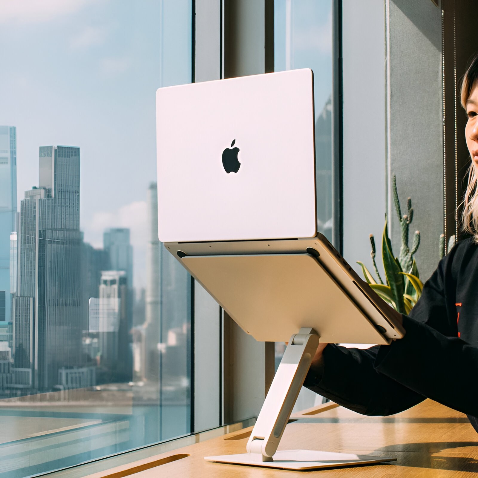 Eine Frau arbeitet an einem MacBook auf einem Ständer vor einem großen Fenster mit Stadtblick.