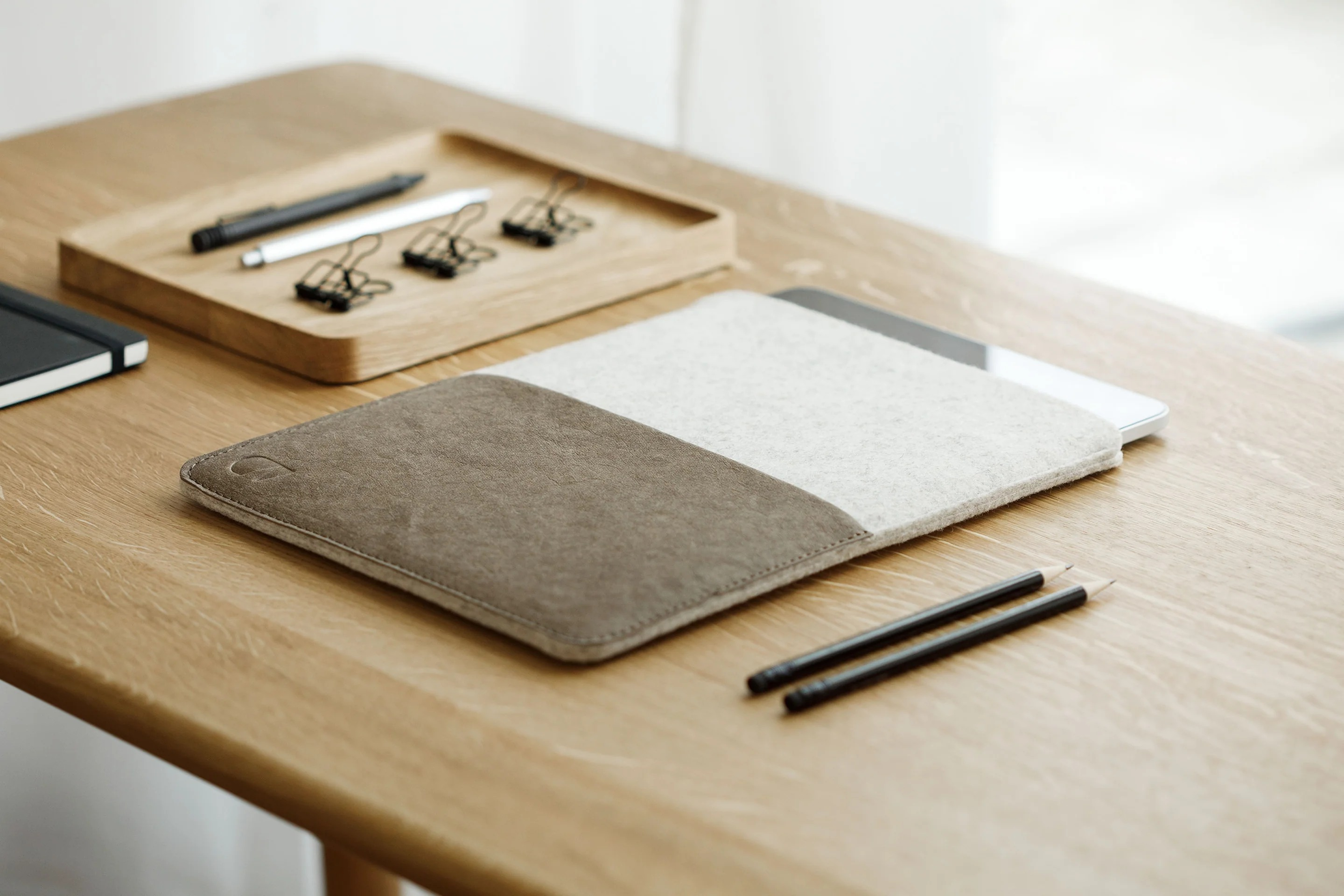 Tablet w szarym filcowo-skórzanym etui na drewnianym biurku, obok leżą ołówki.